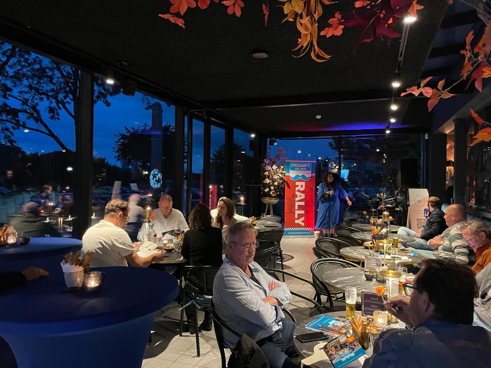 Mensen zitten aan tafels in een felverlichte bar met uitzicht naar buiten in de schemering; iemand staat naast een spandoek met de tekst 