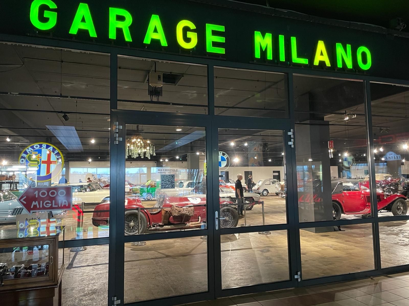 Garage Milano-etalage met klassieke auto's in de etalage. Groen verlicht reclamebord boven de glazen deuren.