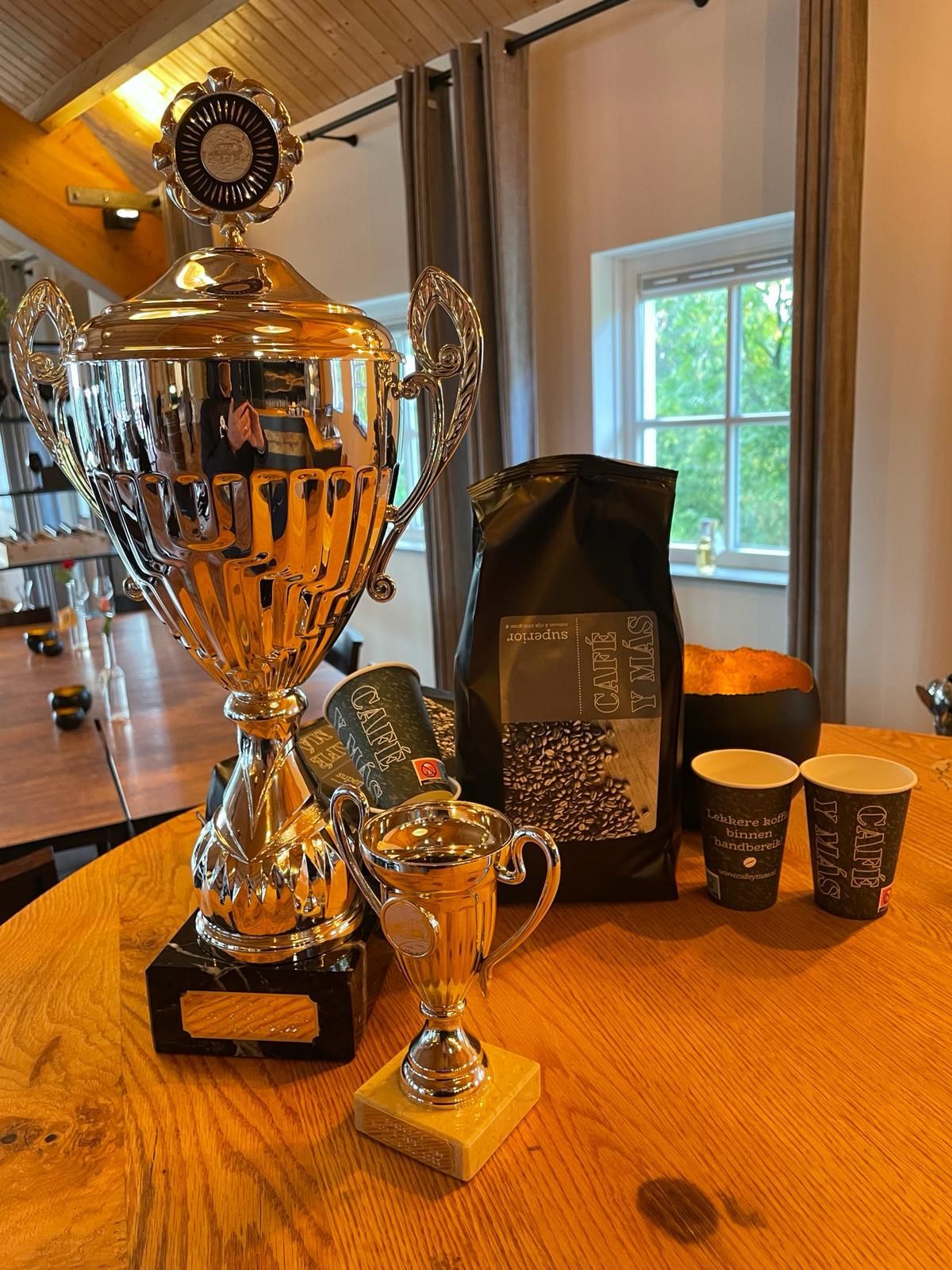 Zilveren trofeeën, een koffiezak en kopjes op een houten tafel, mogelijk gerelateerd aan een koffiewedstrijd.