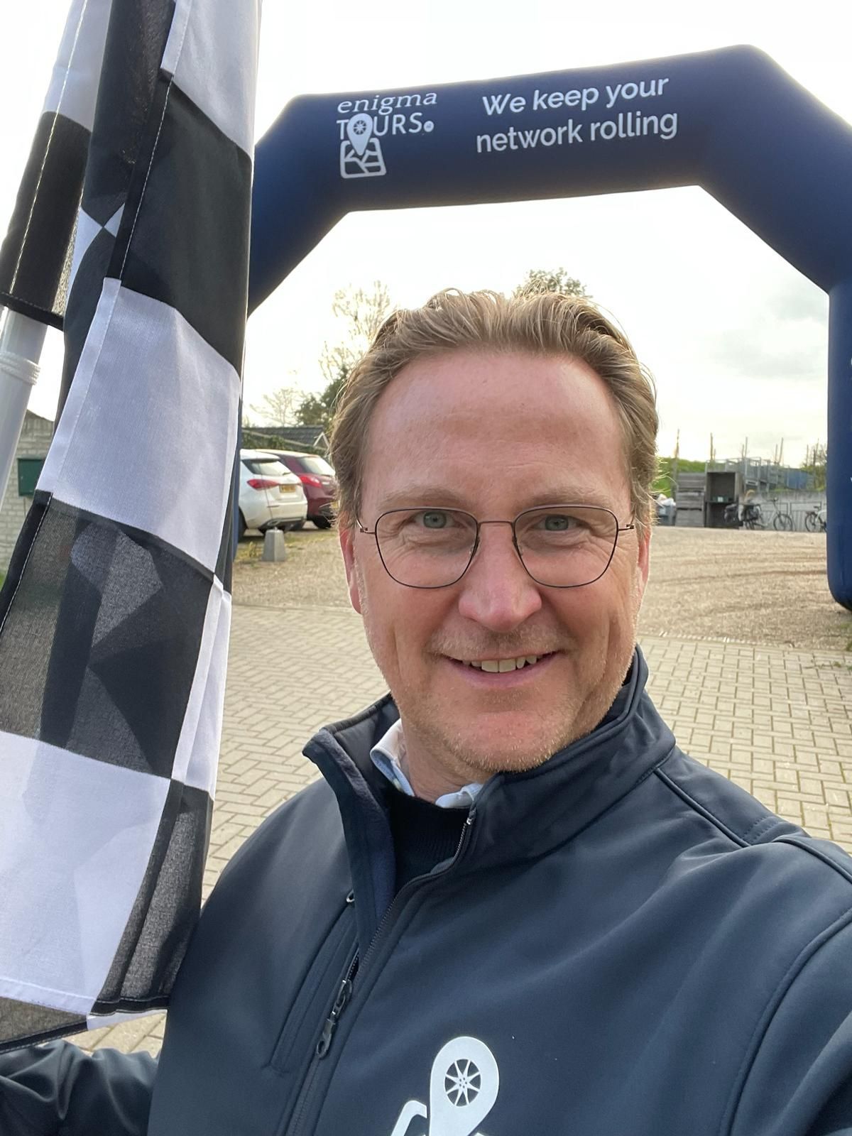 Een man met een geblokte vlag staat lachend voor een boog met de tekst: 