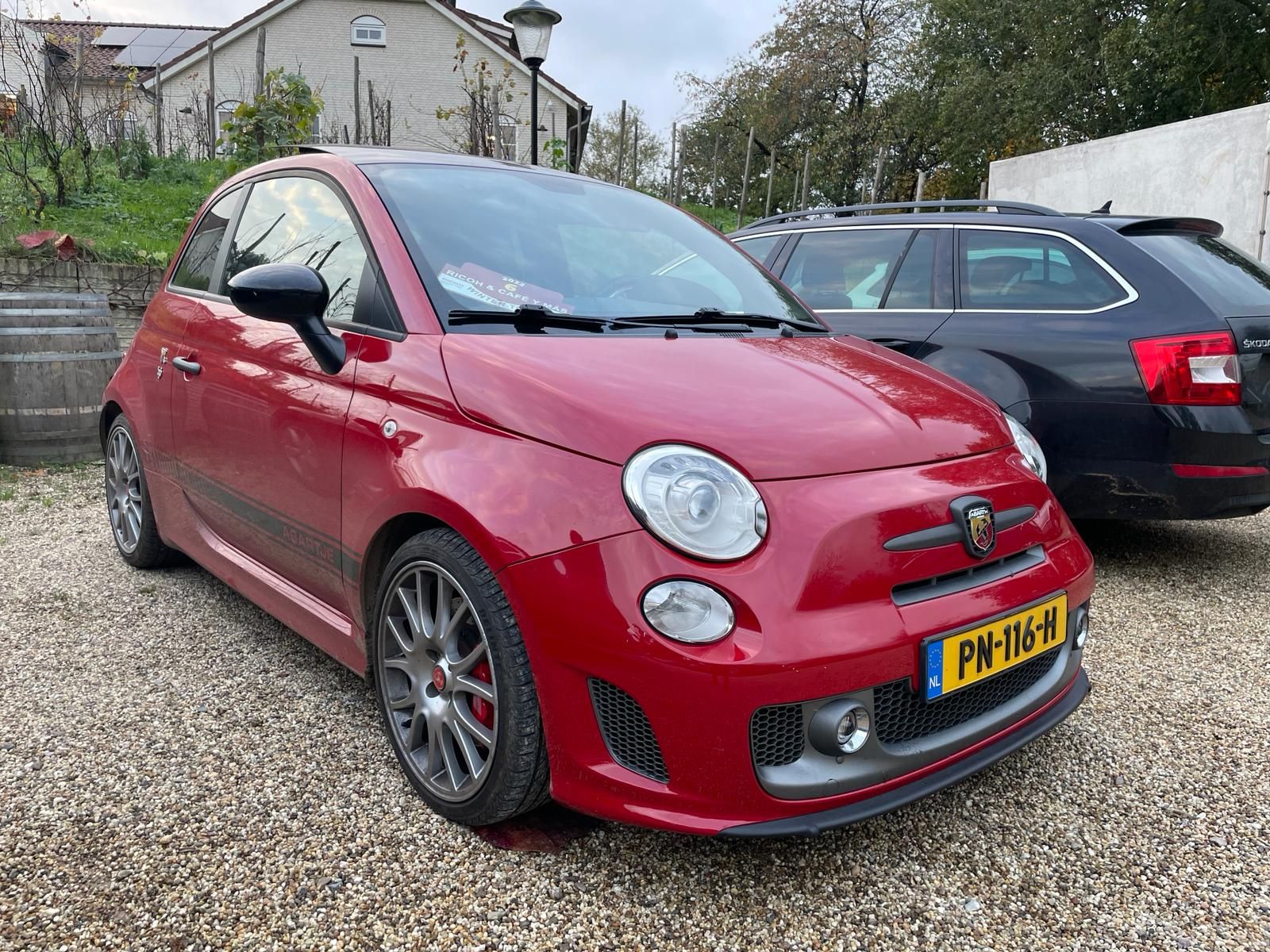 Rode Abarth 500 geparkeerd op grind, met daarachter een donkere auto. Nederlands kenteken.