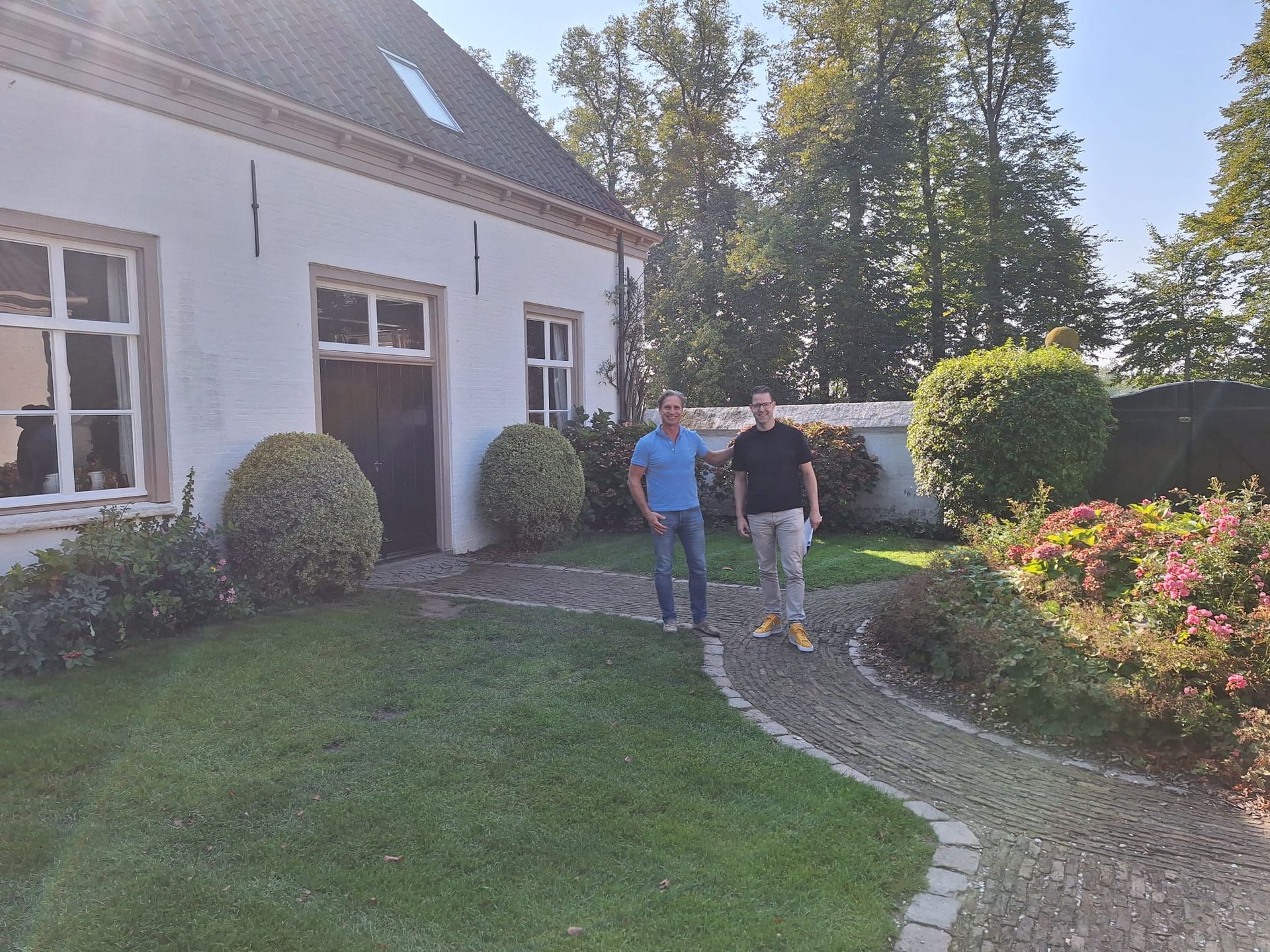 Twee mannen staan voor een wit gebouw met een open deur. Op de voorgrond zijn een groen gazon en struiken te zien.