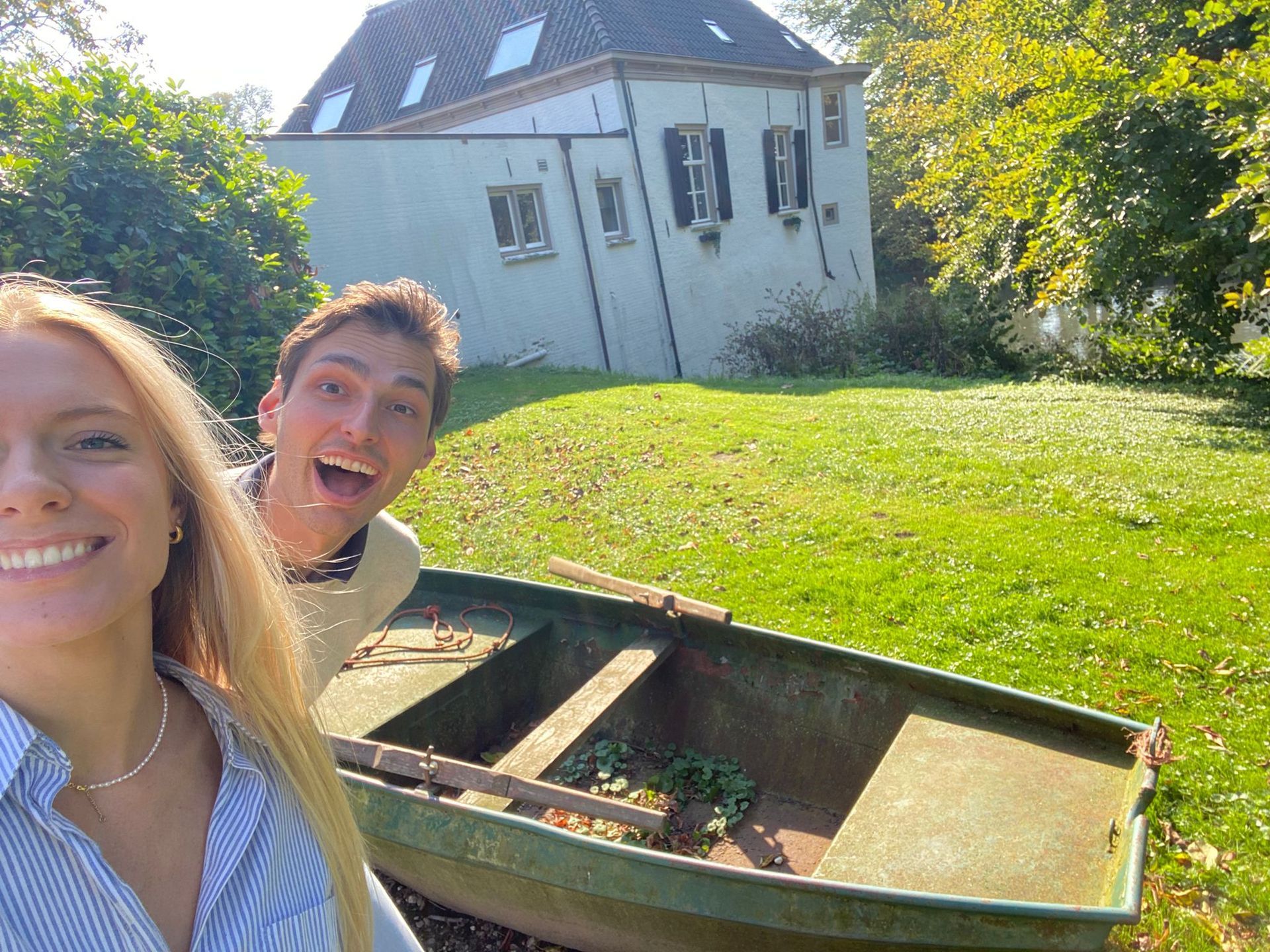 Twee mensen glimlachen bij een oude boot op een grashelling, met een wit huis op de achtergrond.