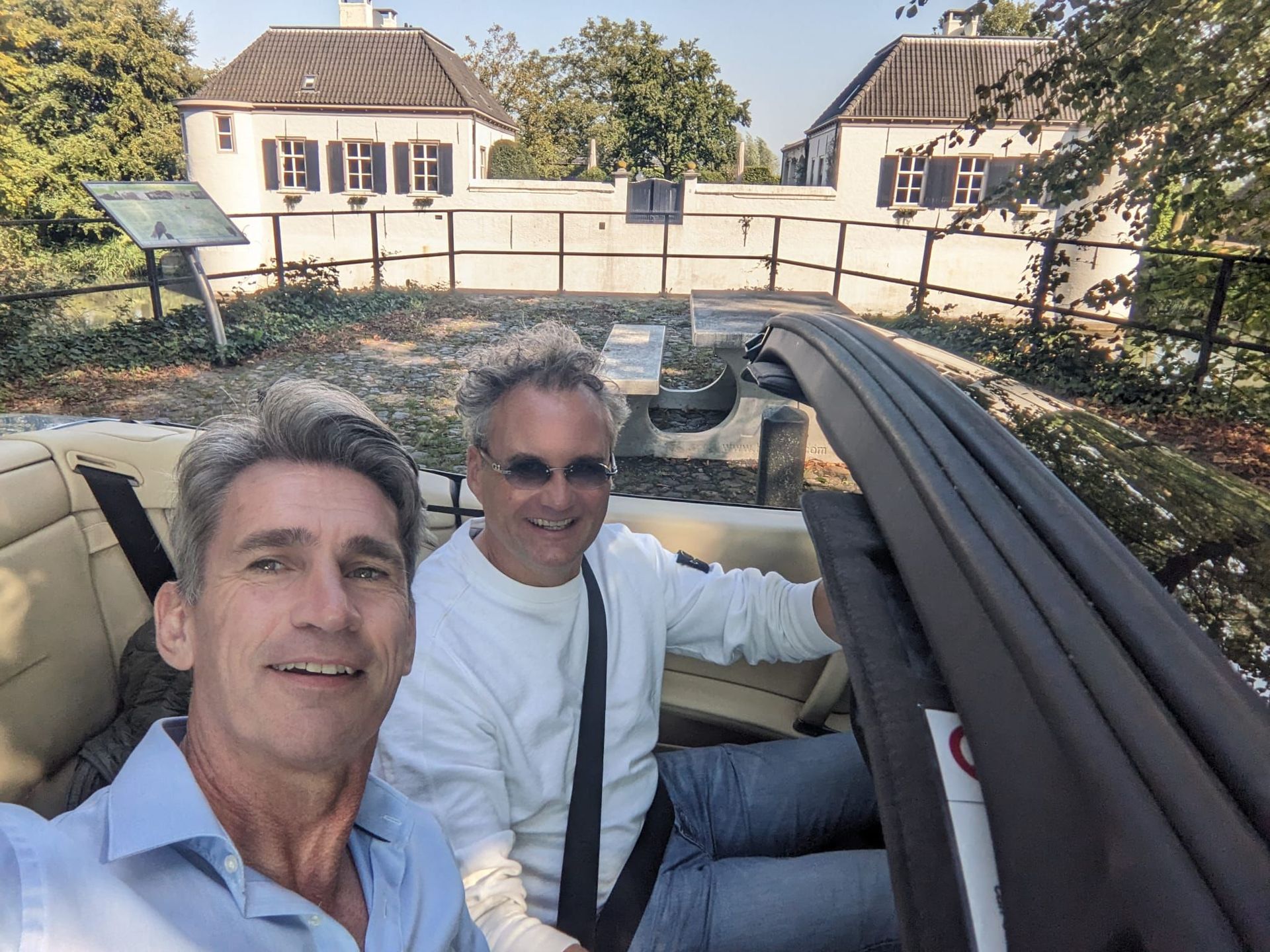 Twee mannen in een cabriolet maken een selfie. Witte gebouwen en groen op de achtergrond. Zonnige dag.