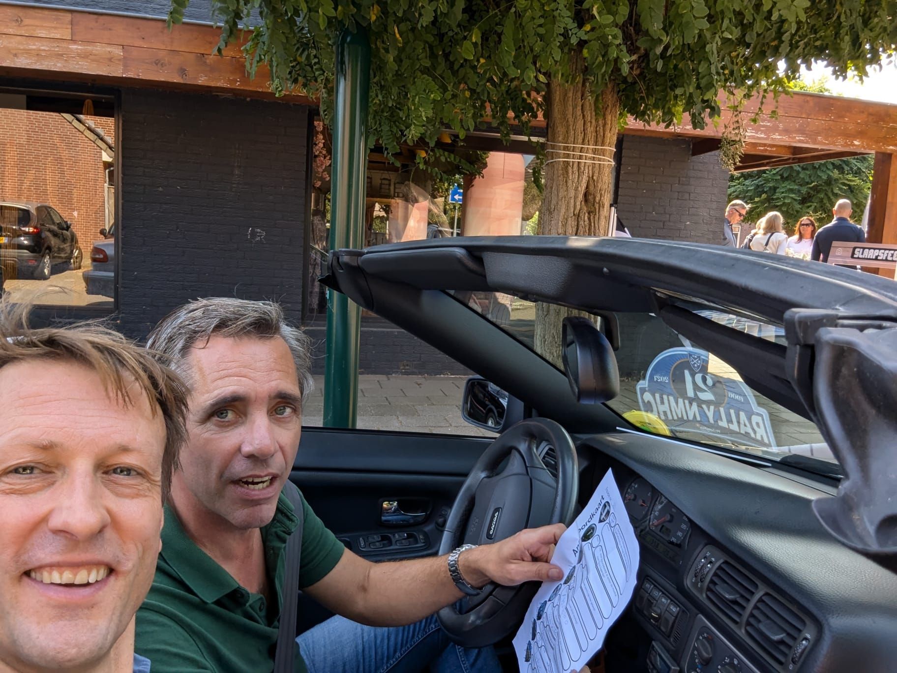 Twee mannen in een cabriolet, één achter het stuur en met papieren in zijn hand, glimlachend. Buiten, zonnige omgeving.