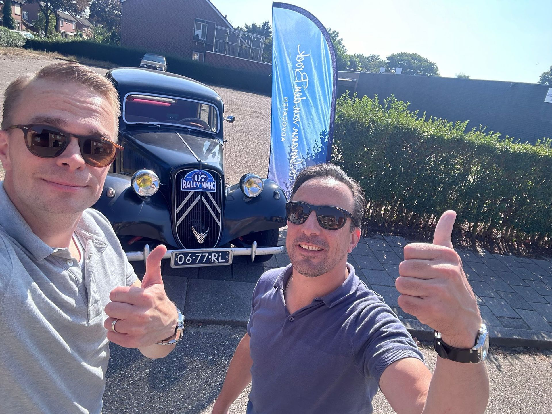 Twee mannen steken hun duim omhoog naast een klassieke zwarte auto met een blauwe vlag op de achtergrond.