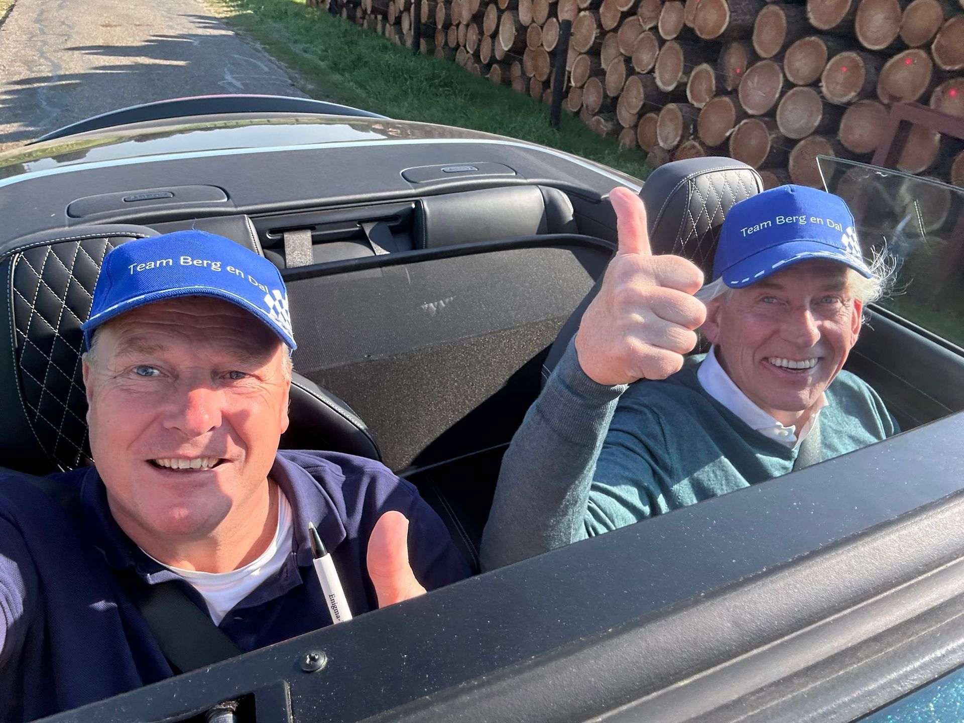 Twee mannen in een cabriolet, met hun duimen omhoog, blauwe petten op en een glimlach op hun gezicht.