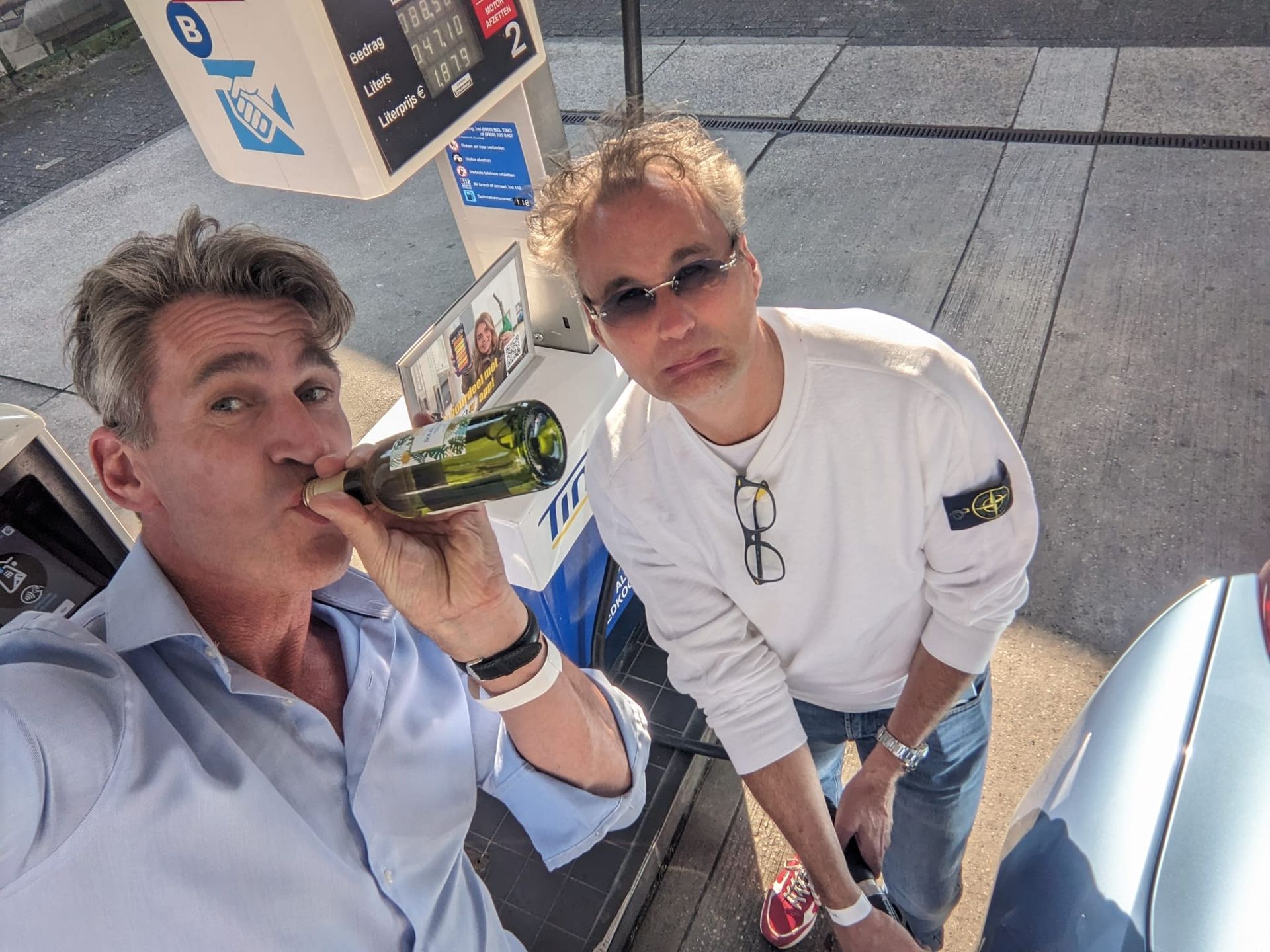 Twee mannen bij een benzinepomp; de ene drinkt uit een fles, de andere houdt een brandstofslang vast.