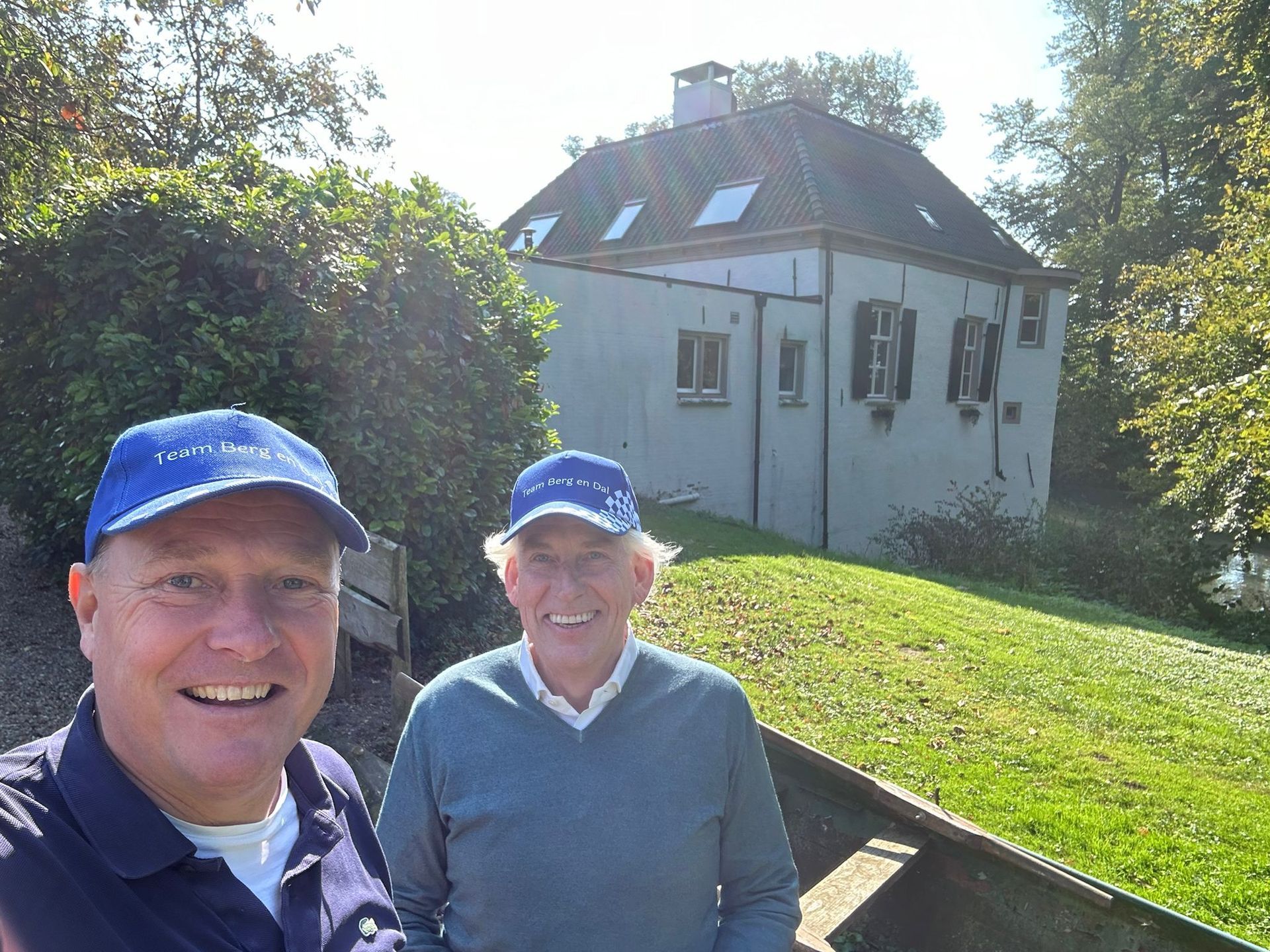 Twee mannen met blauwe hoeden glimlachen terwijl ze poseren voor een lichtgekleurd gebouw met een schuin dak, een groen gazon en bomen.