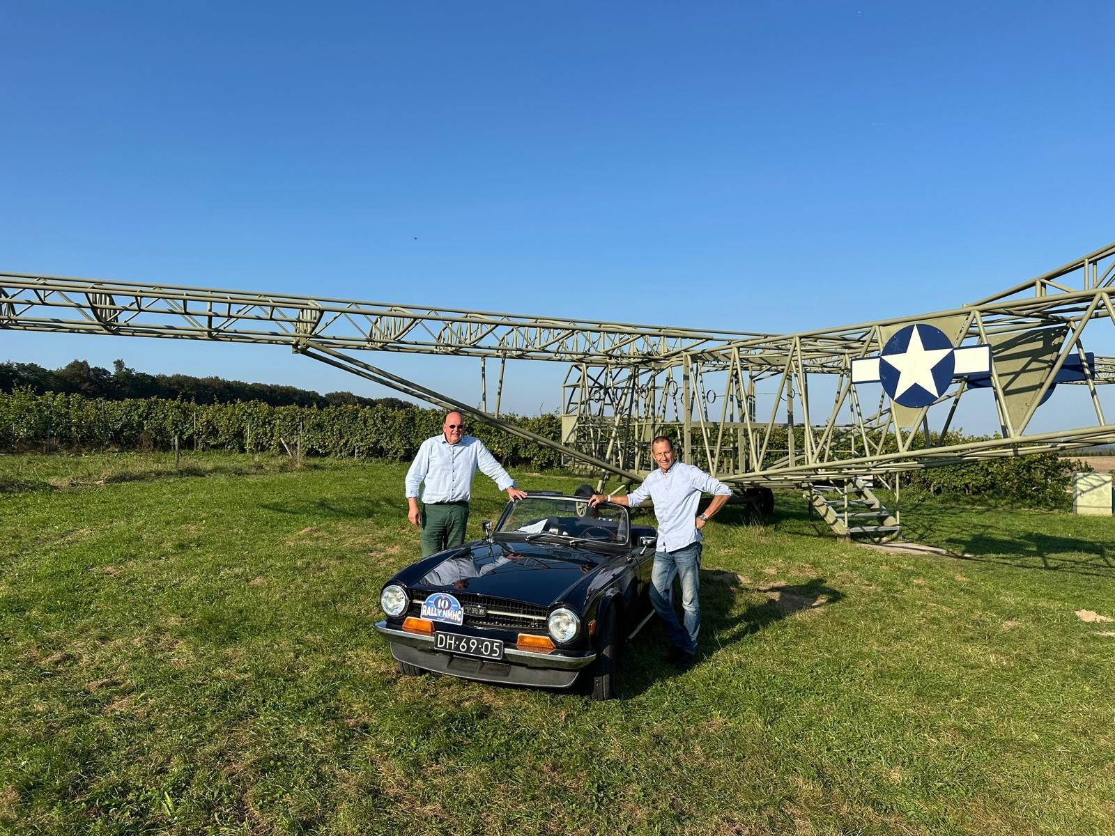 Twee mannen staan naast een zwarte cabriolet op een grasveld, met op de achtergrond een metalen vliegtuigconstructie.