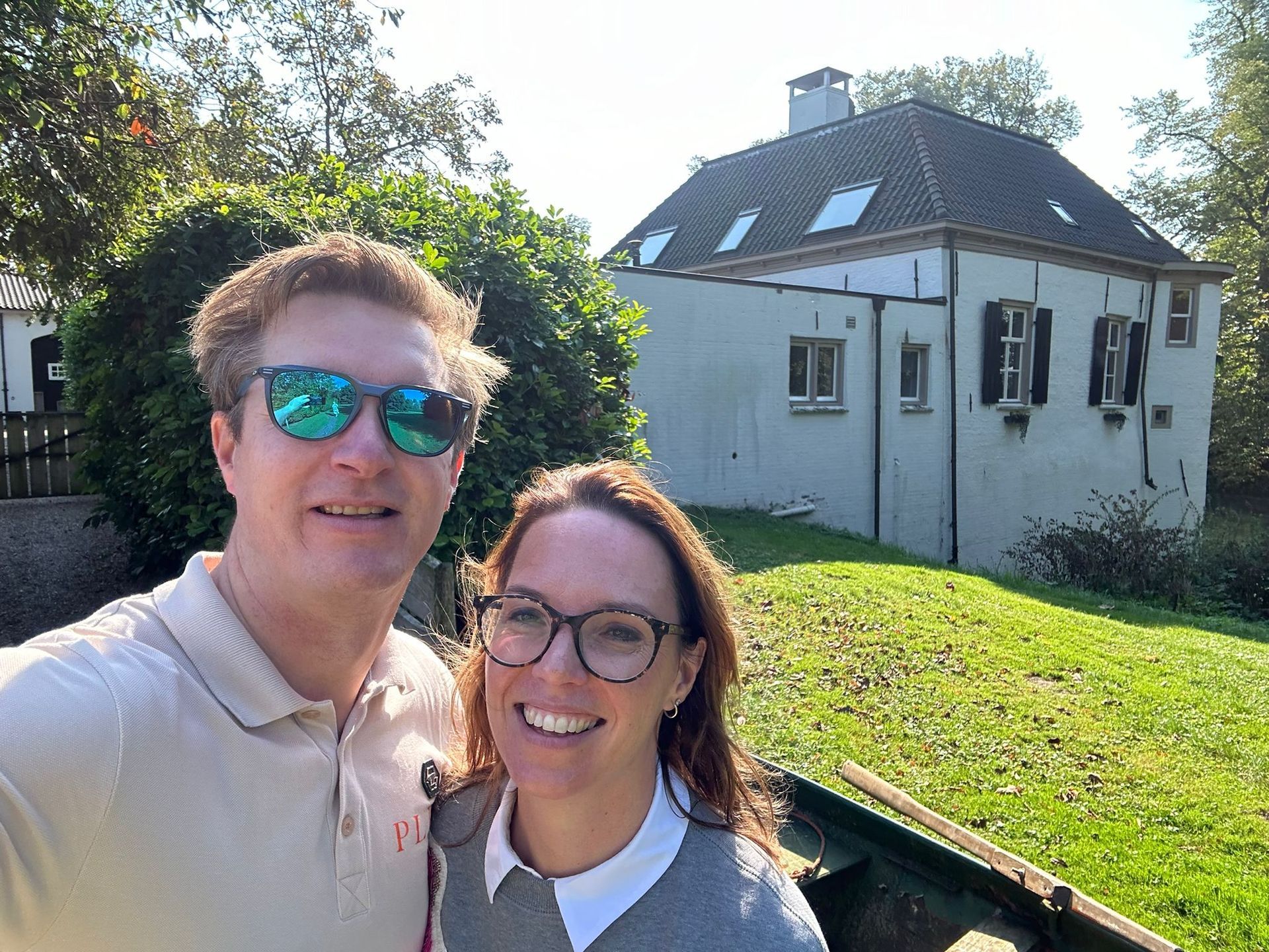 Een lachend stel maakt een selfie bij een lichtgekleurd huis met een donker dak, op een zonnige dag.