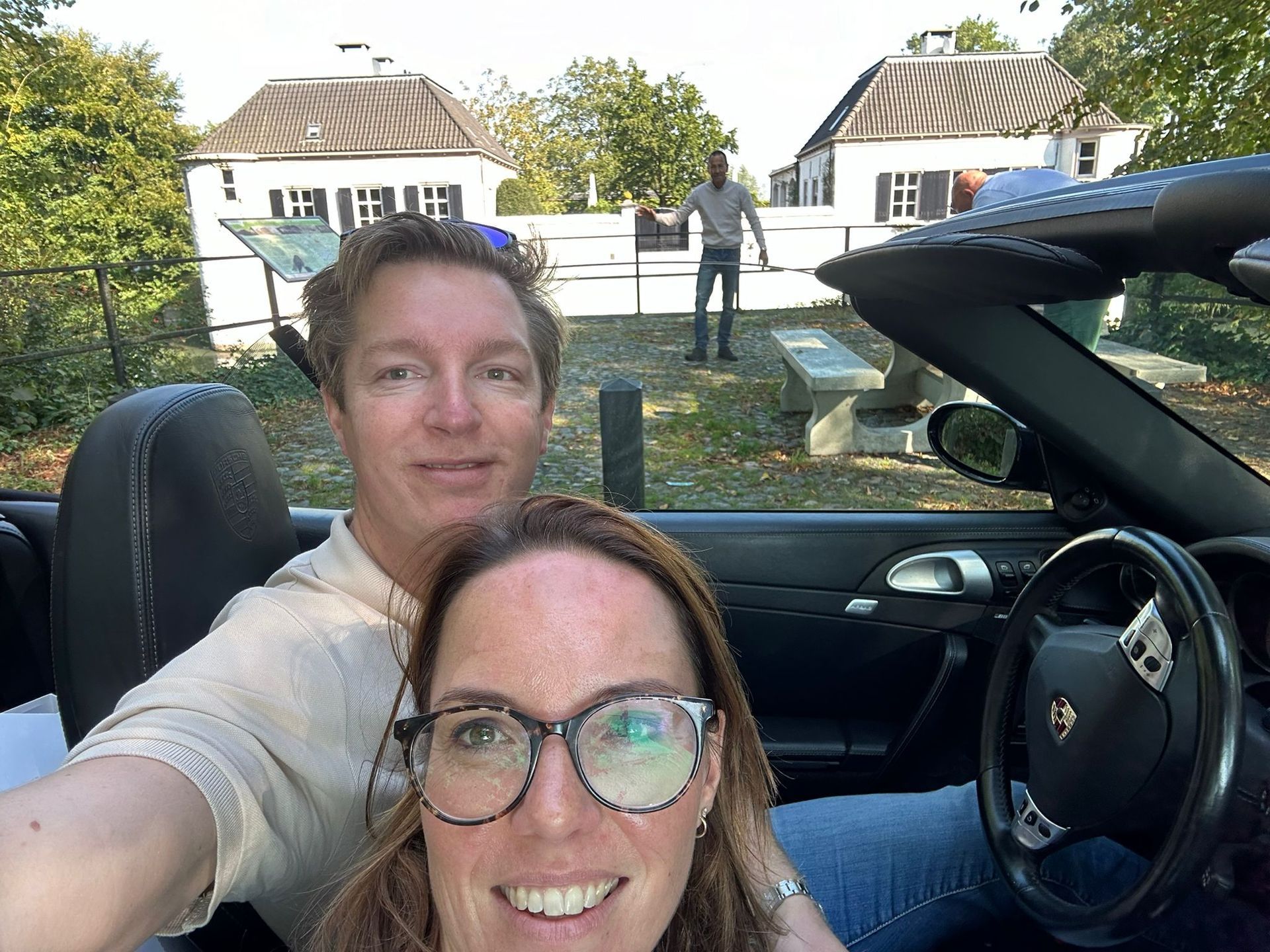 Een stel in een cabriolet, lachend, met een gebouw op de achtergrond, op een zonnige dag.