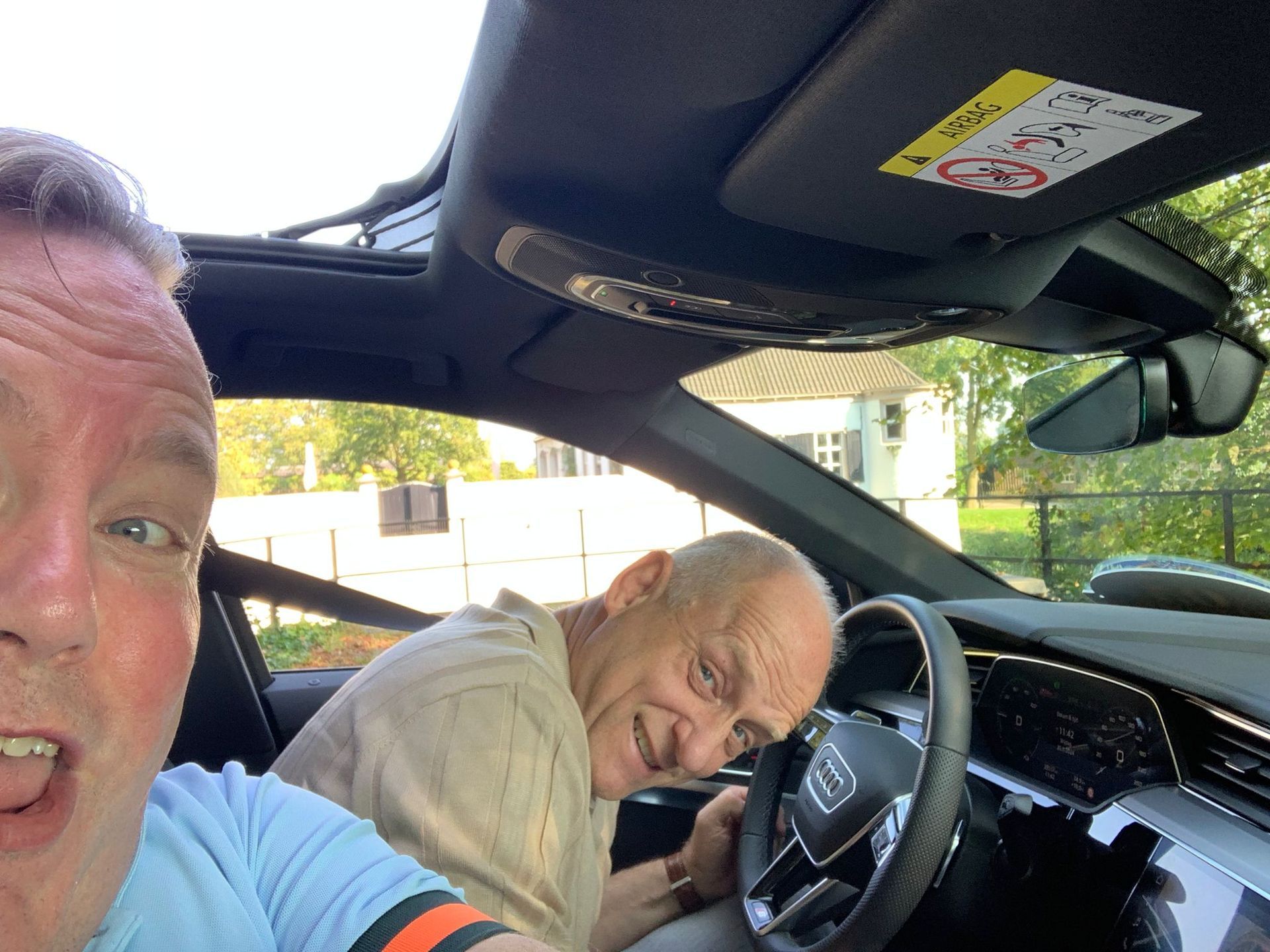 Twee mannen in een auto; de ene maakt een selfie, de andere lacht naar het stuur. Binnen in de auto, open schuifdak.
