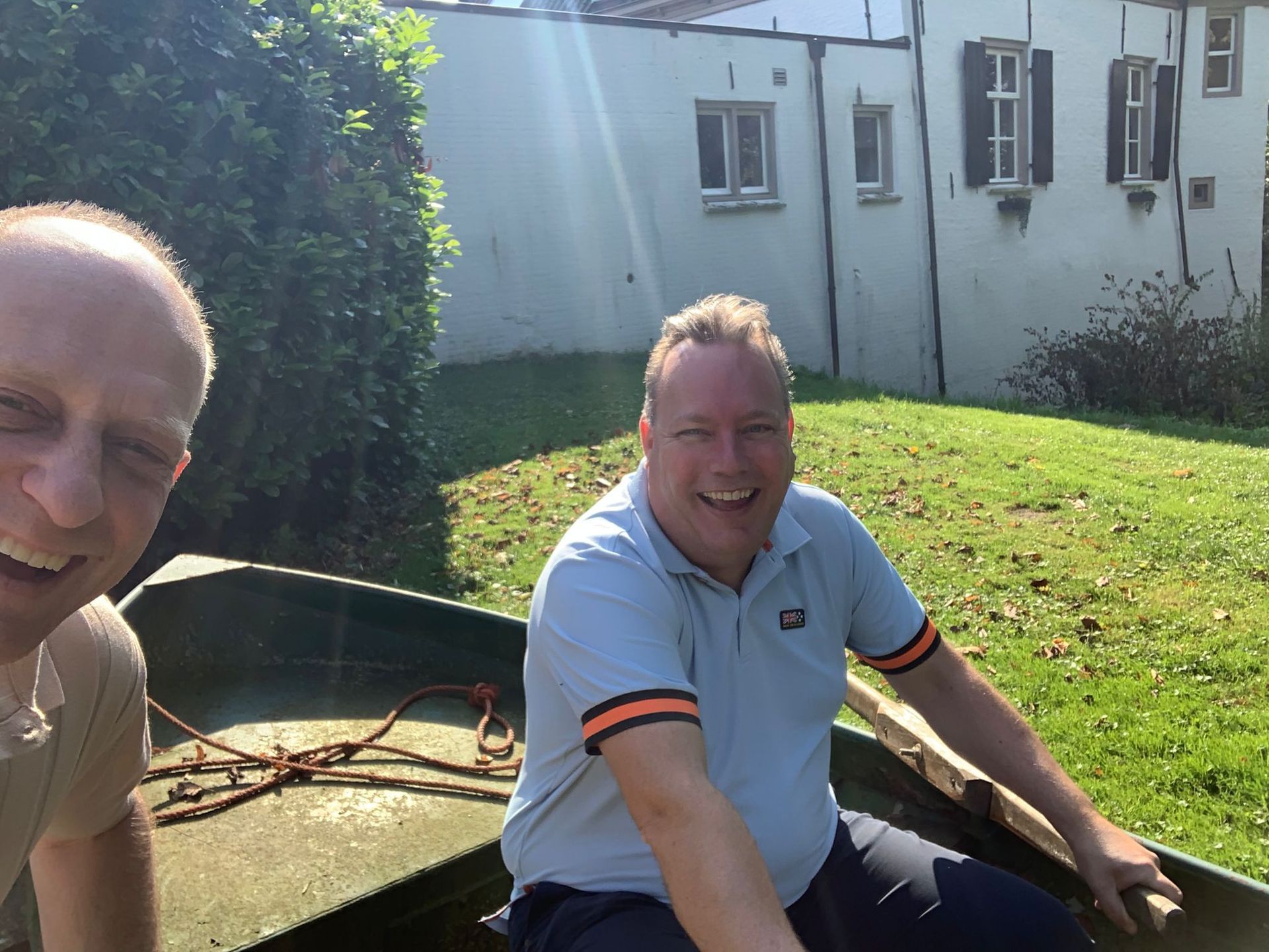 Twee mannen glimlachen in een klein bootje op een zonnige dag, vlakbij een wit gebouw met groen gras.