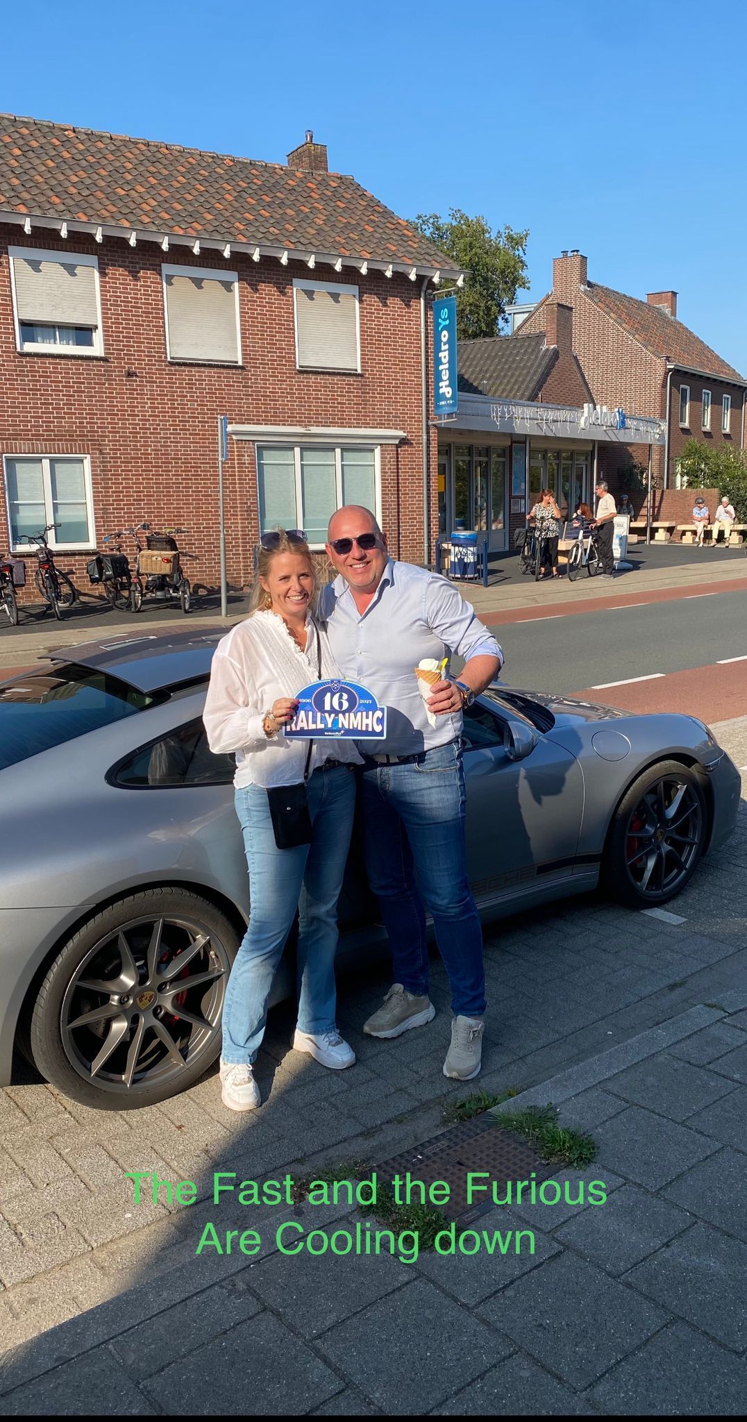 Een stel staat naast een zilverkleurige sportwagen met een taart in hun handen. Ze bevinden zich buiten, vlakbij een bakstenen gebouw.