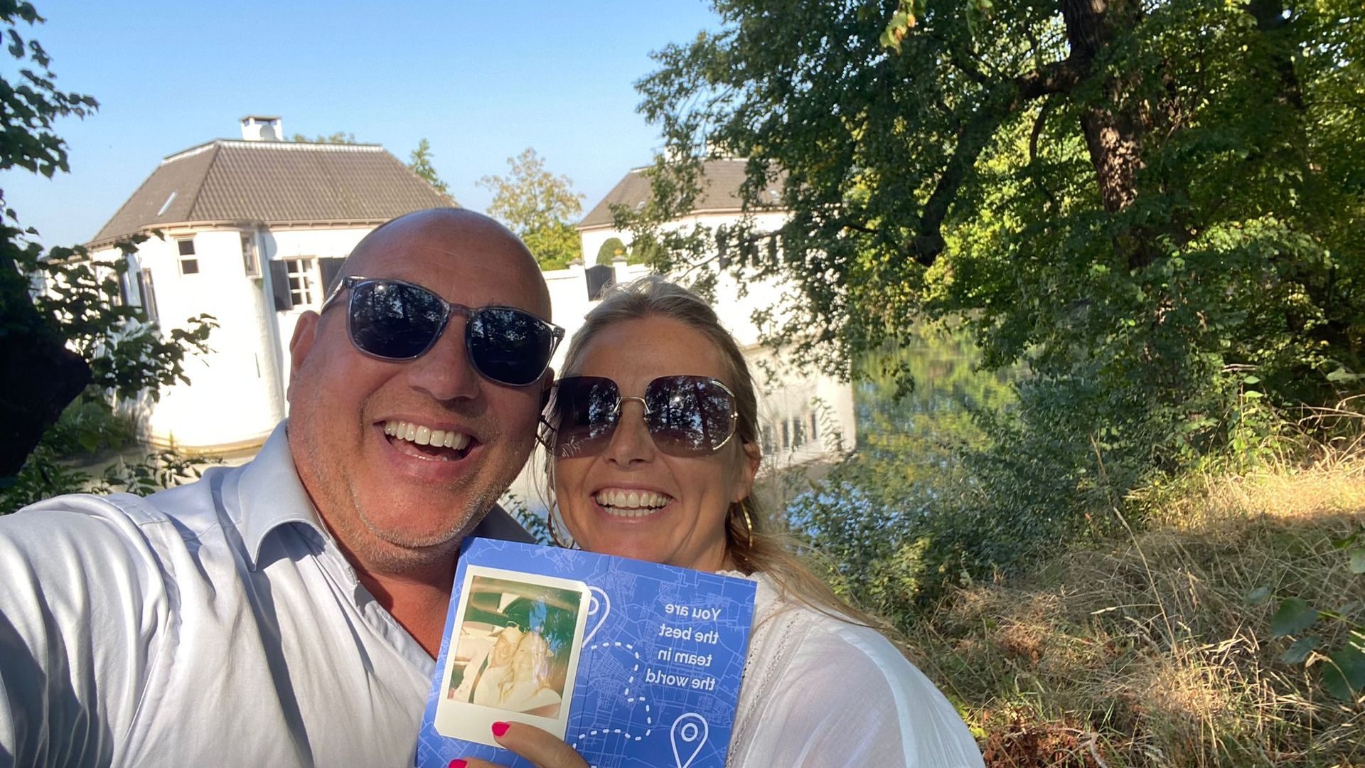 Een man en een vrouw glimlachen en houden een blauw boek vast. Op de achtergrond zijn een wit gebouw en bomen te zien. Een zonnige buitenomgeving.