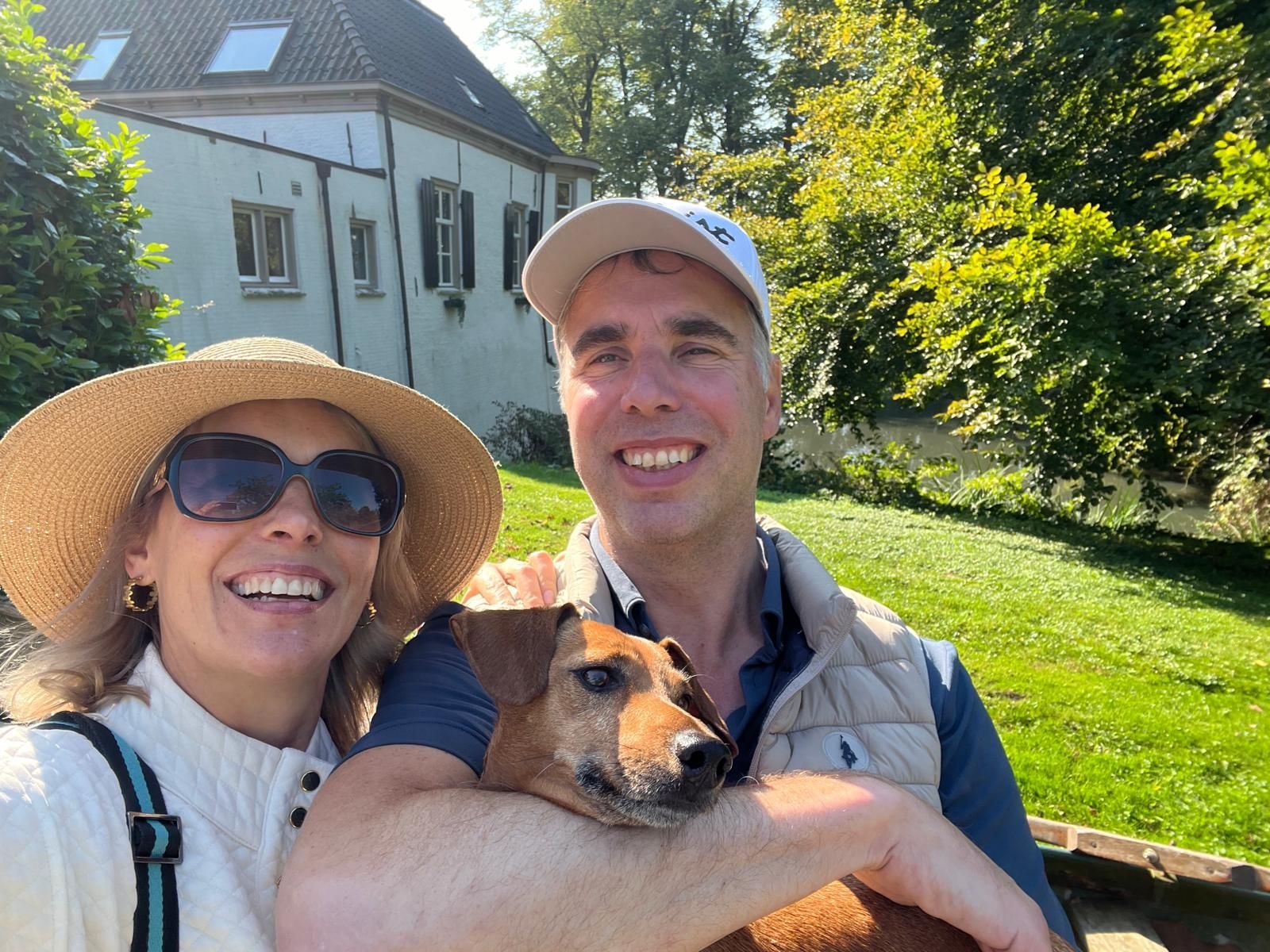 Een stel met een hond lacht buiten voor een gebouw en bomen op een zonnige dag.