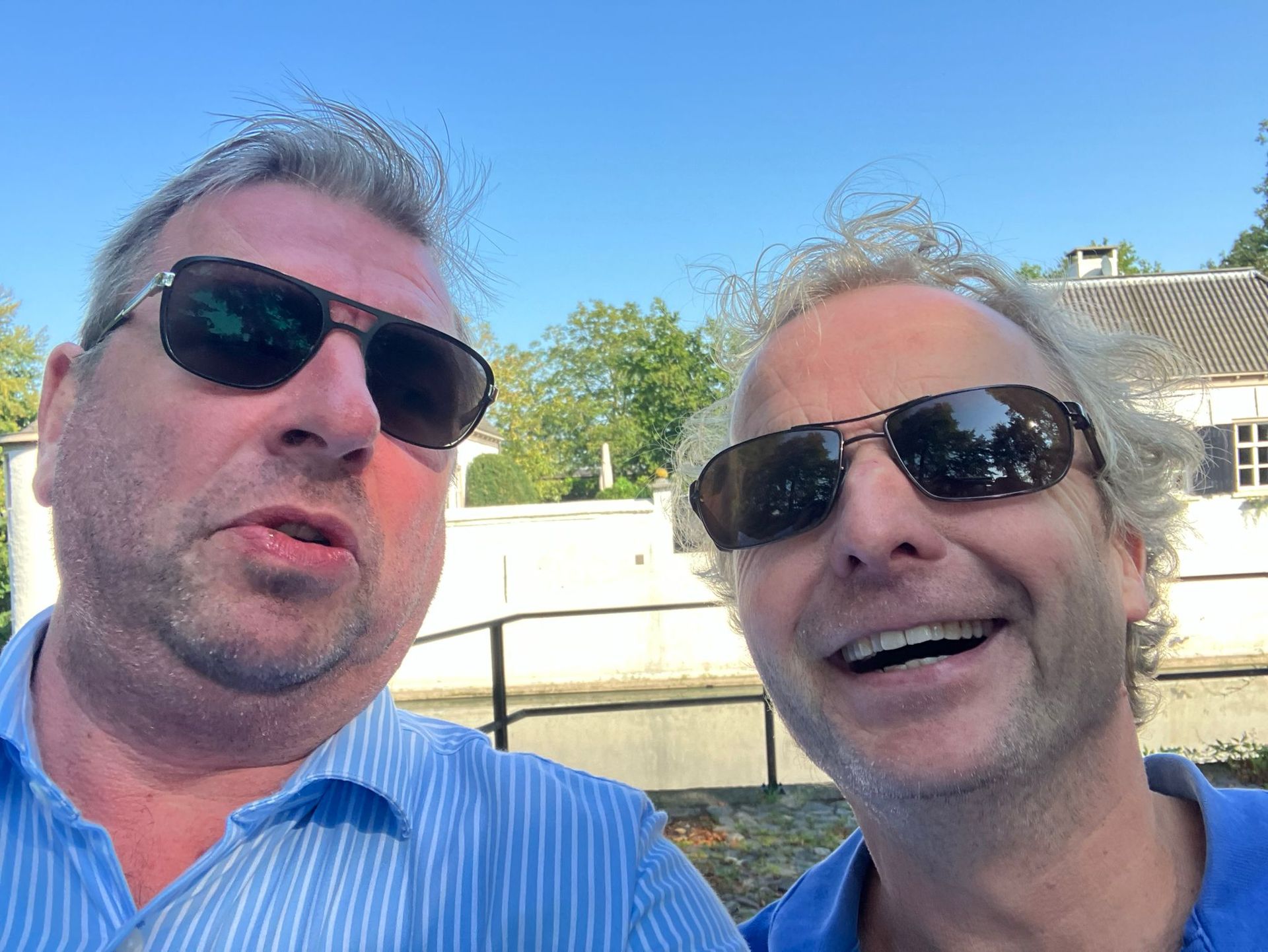 Twee mannen met zonnebrillen maken een selfie in de buitenlucht, vlakbij een wateroppervlak, en lachen.