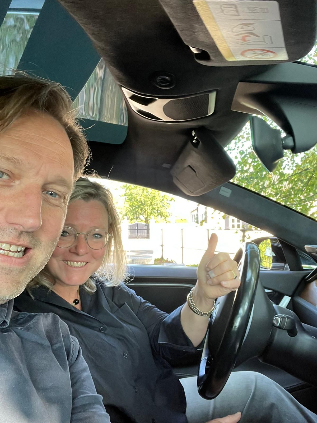 Een man en een vrouw zitten in een auto; de vrouw steekt haar duim omhoog. Ze zitten binnen, in een donker interieur met zonlicht.
