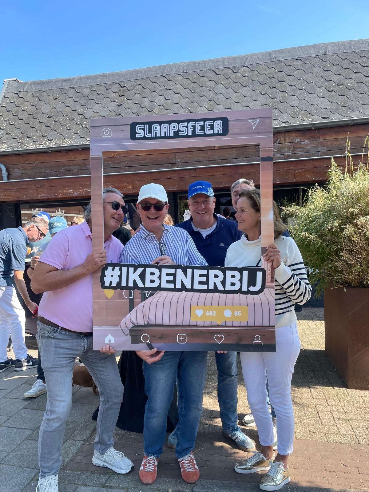 Vier mensen poseren met een fotolijstje tijdens een evenement in de buitenlucht. Op het lijstje staat