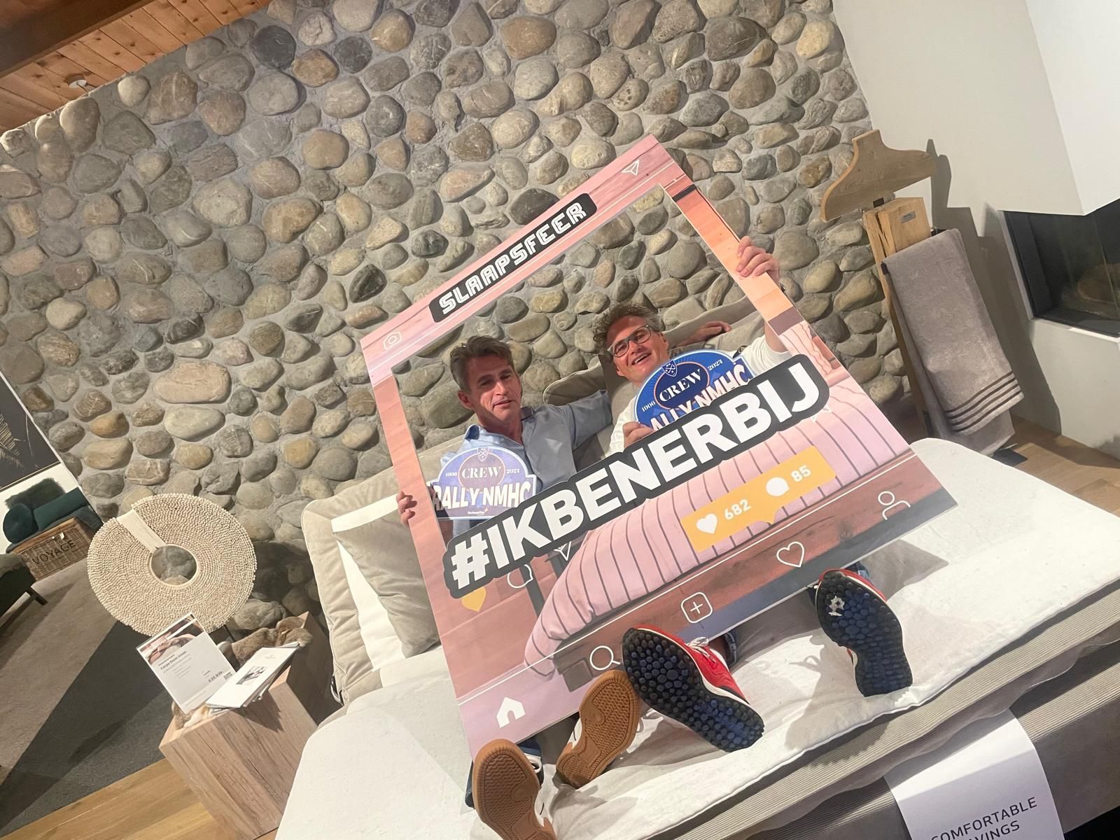 Twee mensen poseren achter een kartonnen frame, op een bed voor een stenen muur. Schoenen staan op het bed.