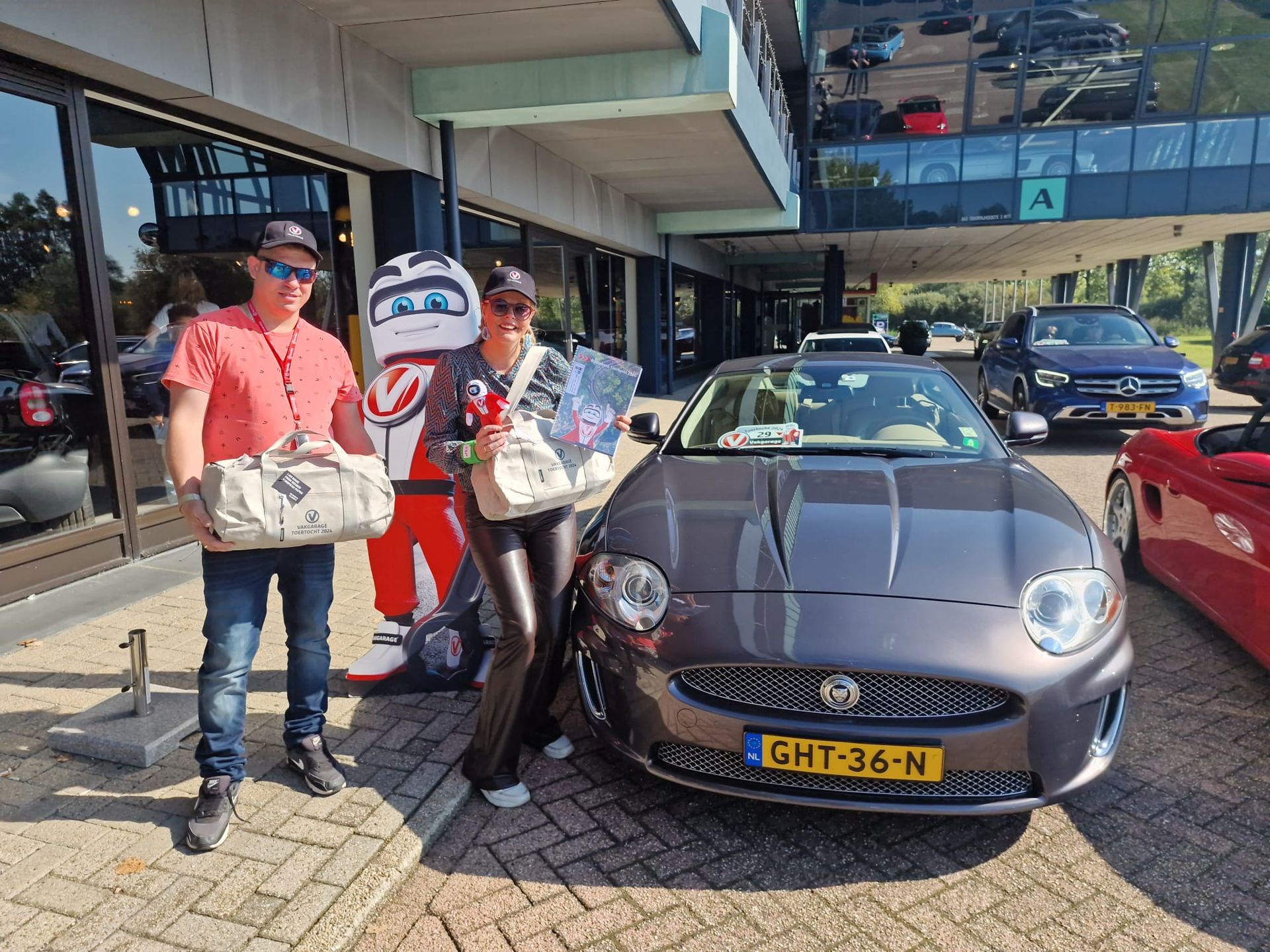 Een man en een vrouw poseren met een Jaguar; een mascotte is op de achtergrond te zien. Buitenopname met auto's en een gebouw.