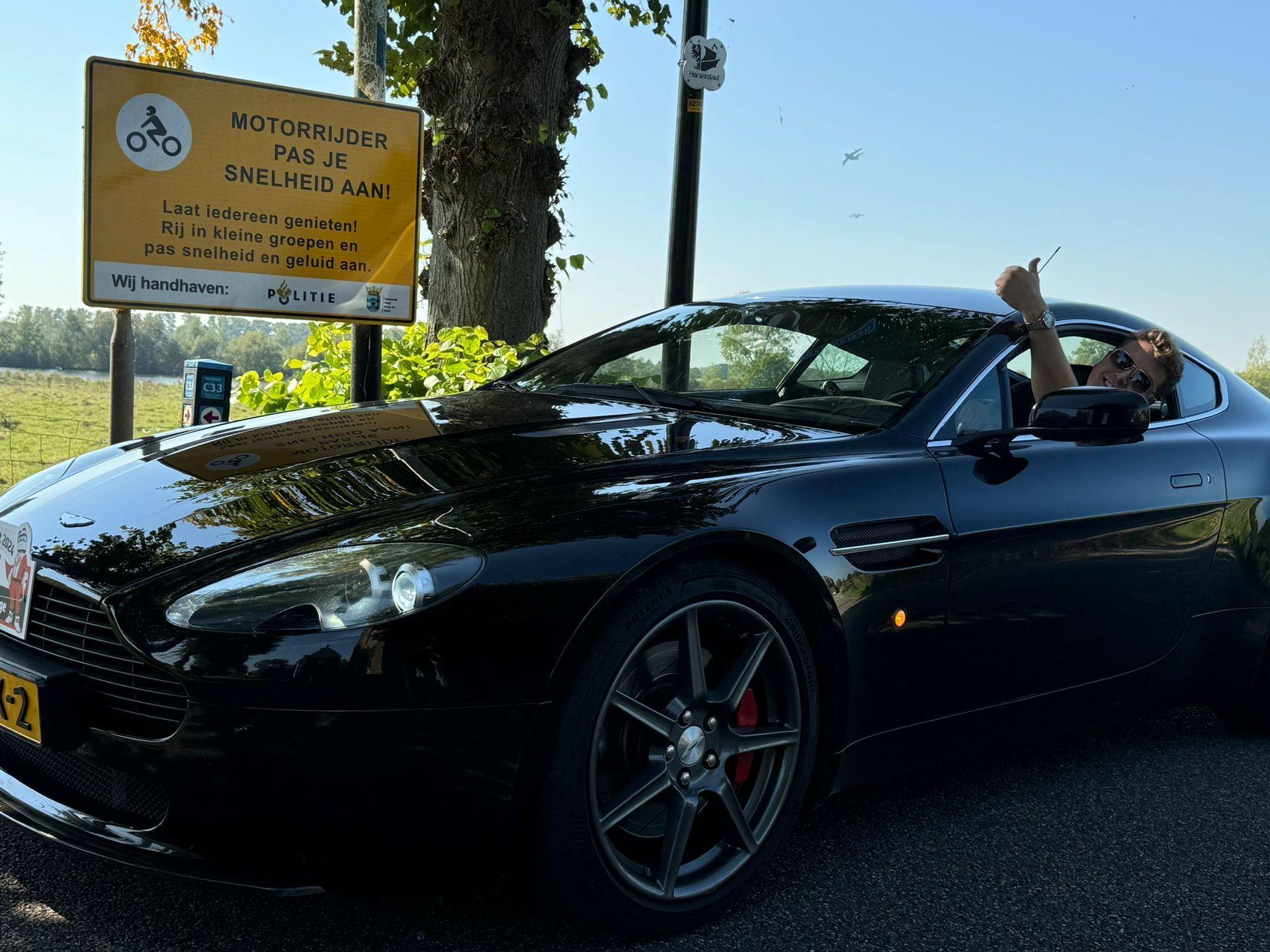 Een zwarte Aston Martin staat geparkeerd bij een bord; er zit iemand achter het stuur, met een opgeheven hand in een gebaar.