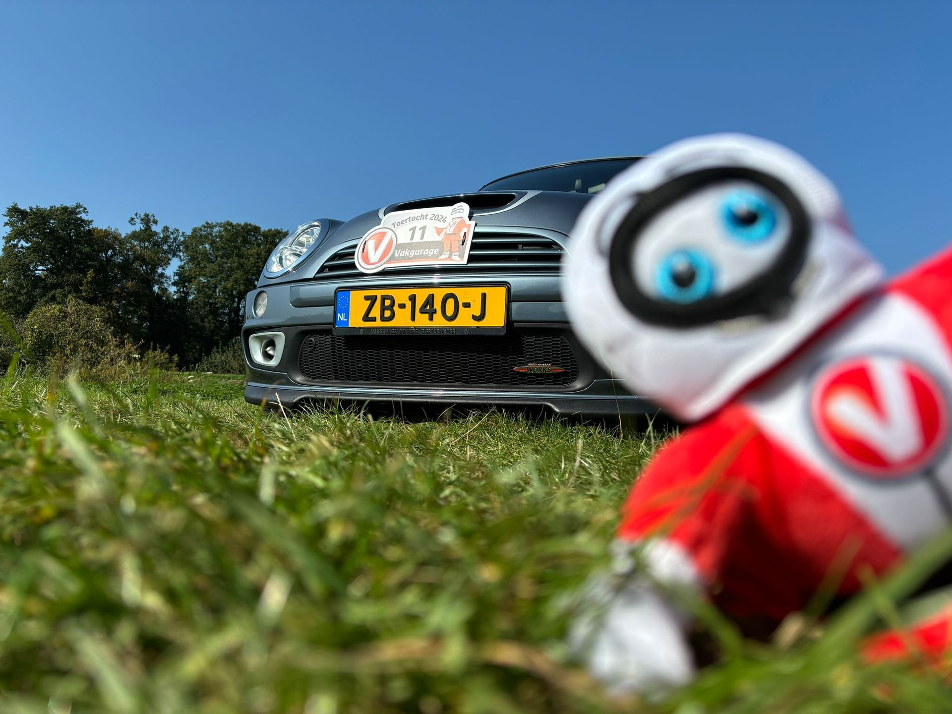 Een knuffelrobot in het gras, tegenover een grijze Mini Cooper met Nederlands kenteken ZB-140-J op een zonnige dag.