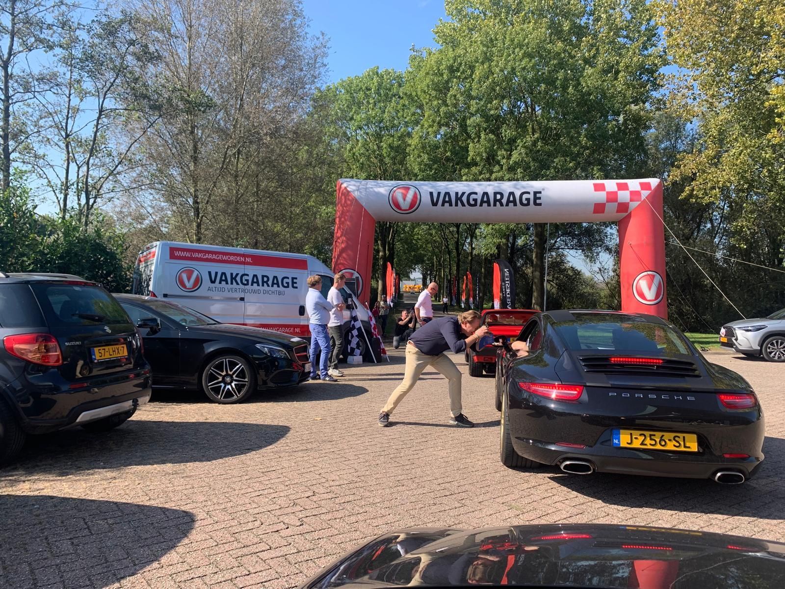 Auto's geparkeerd bij een poort van VANkGARAGE. Mensen verzamelen zich. Een man leunt in een zwarte sportwagen. Een zonnige dag.
