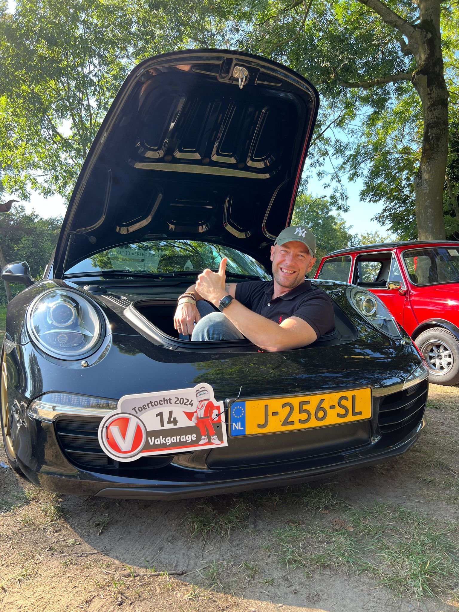 Een man steekt zijn duim omhoog terwijl hij op de open motorkap van een zwarte Porsche zit.