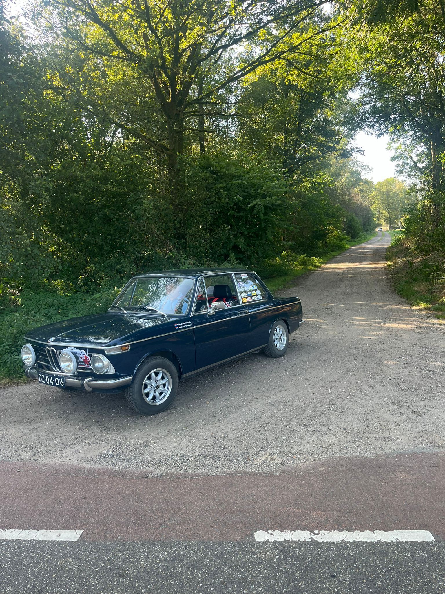 Donkerblauwe oldtimer geparkeerd op een grindweg, omringd door bomen.
