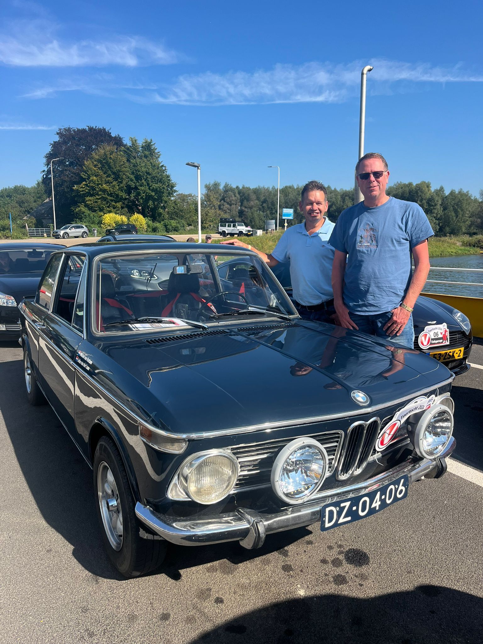 Twee mannen poseren bij een blauwe oldtimer BMW. Buiten, zonnig weer.