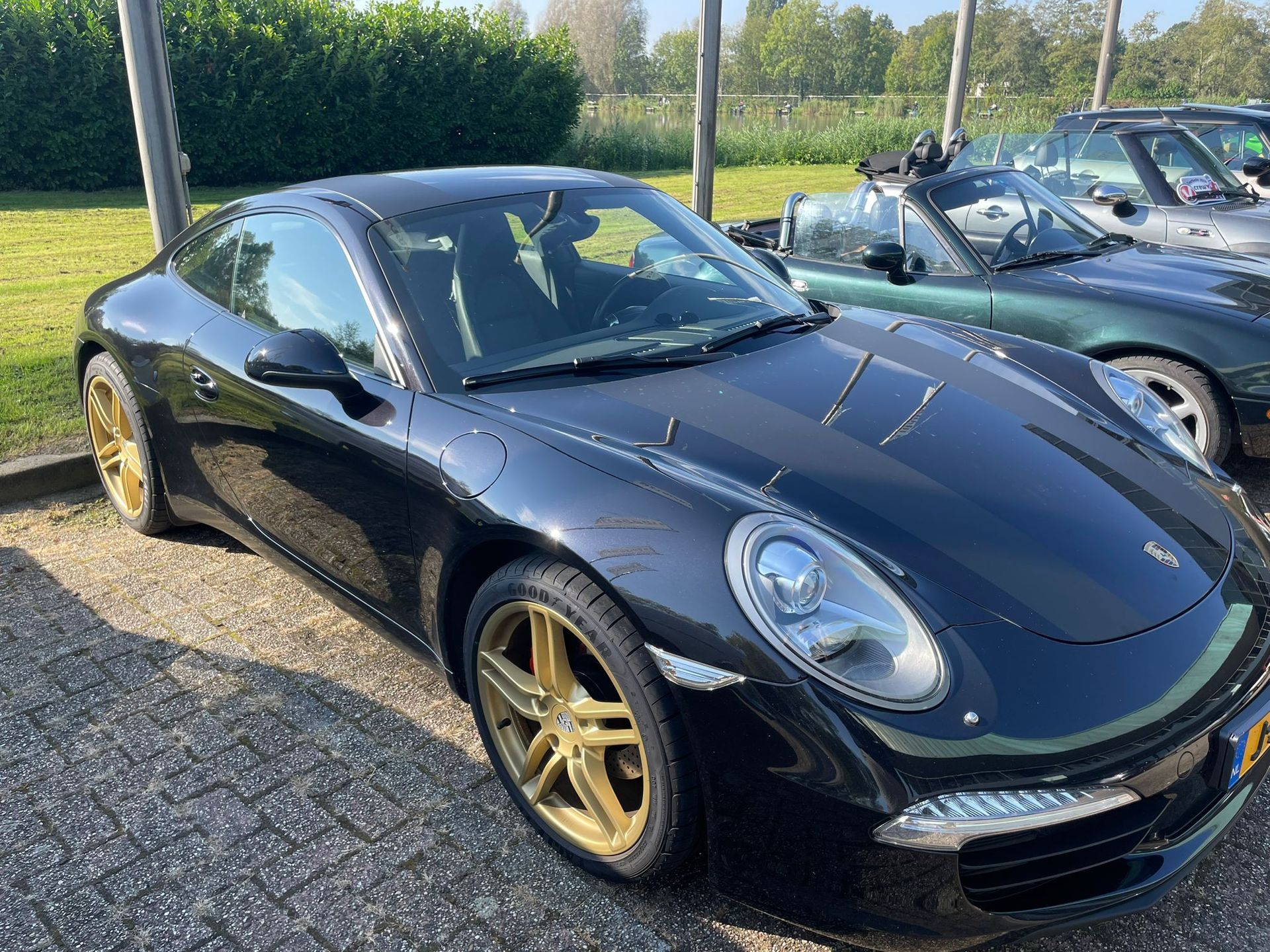 Een zwarte Porsche 911 met gouden velgen staat op een zonnige dag buiten geparkeerd naast een groene cabriolet.