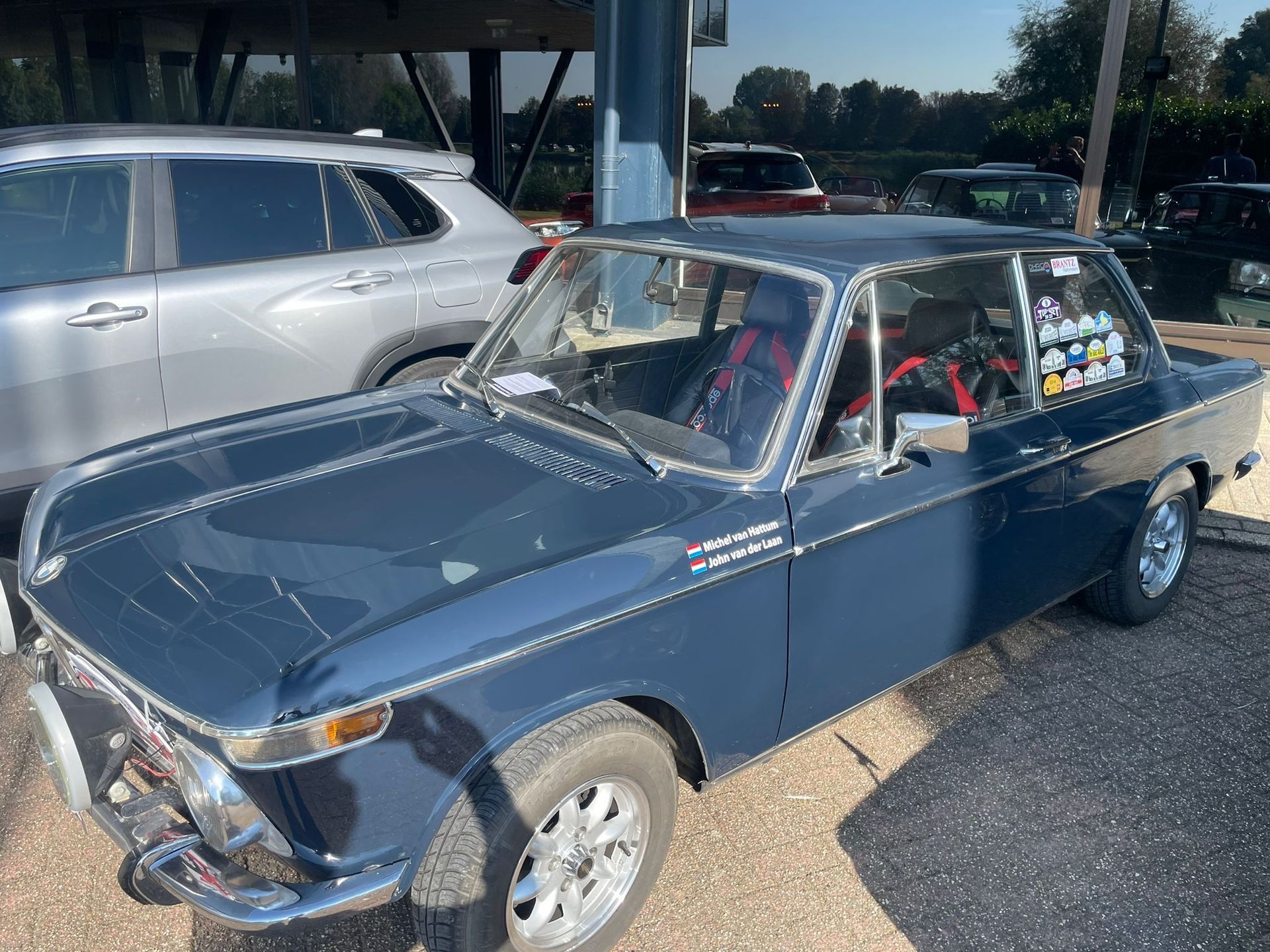 Een blauwe vintage BMW 2002 staat buiten geparkeerd op een zonnige dag. De chromen sierlijsten en de racestoel zijn zichtbaar.