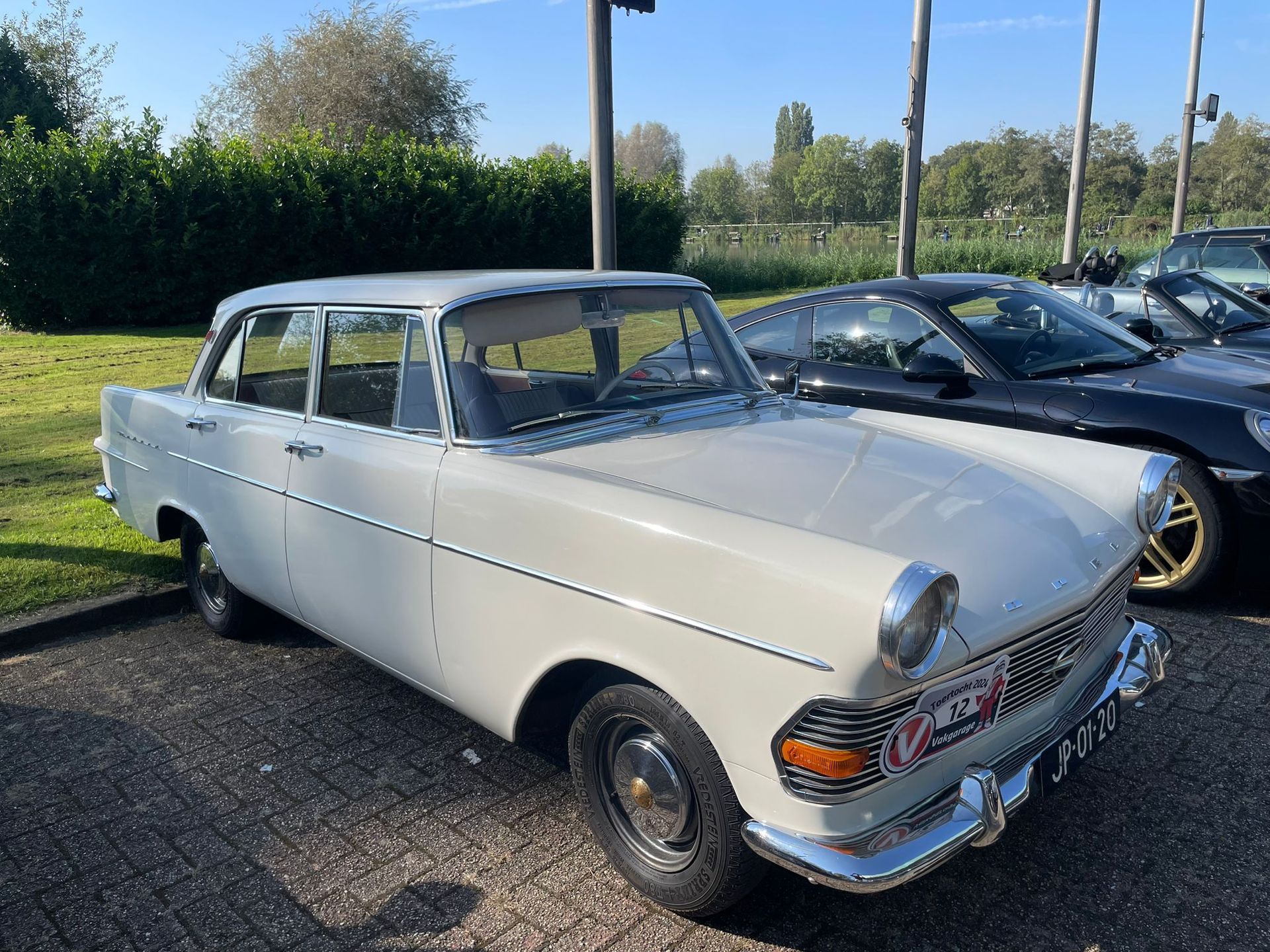 Een vintage lichtgekleurde sedan staat op een zonnige dag buiten geparkeerd, met daarachter een zwarte auto zichtbaar.
