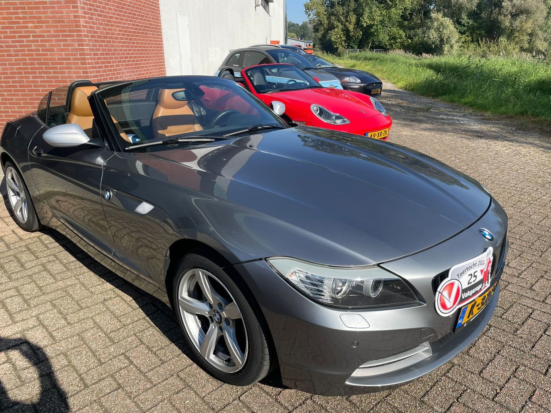Grijze BMW Z4 cabriolet met beige interieur geparkeerd op een verharde weg. Rode auto op de achtergrond.