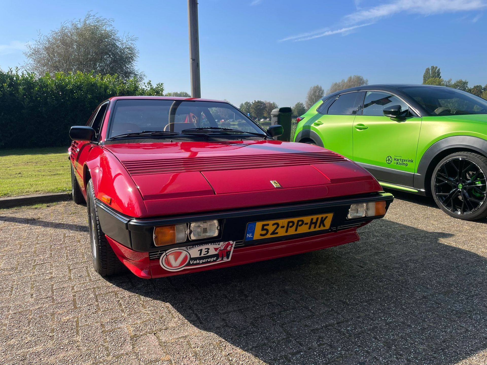 Een rode oldtimer sportwagen met Nederlands kenteken staat geparkeerd naast een groene moderne auto.