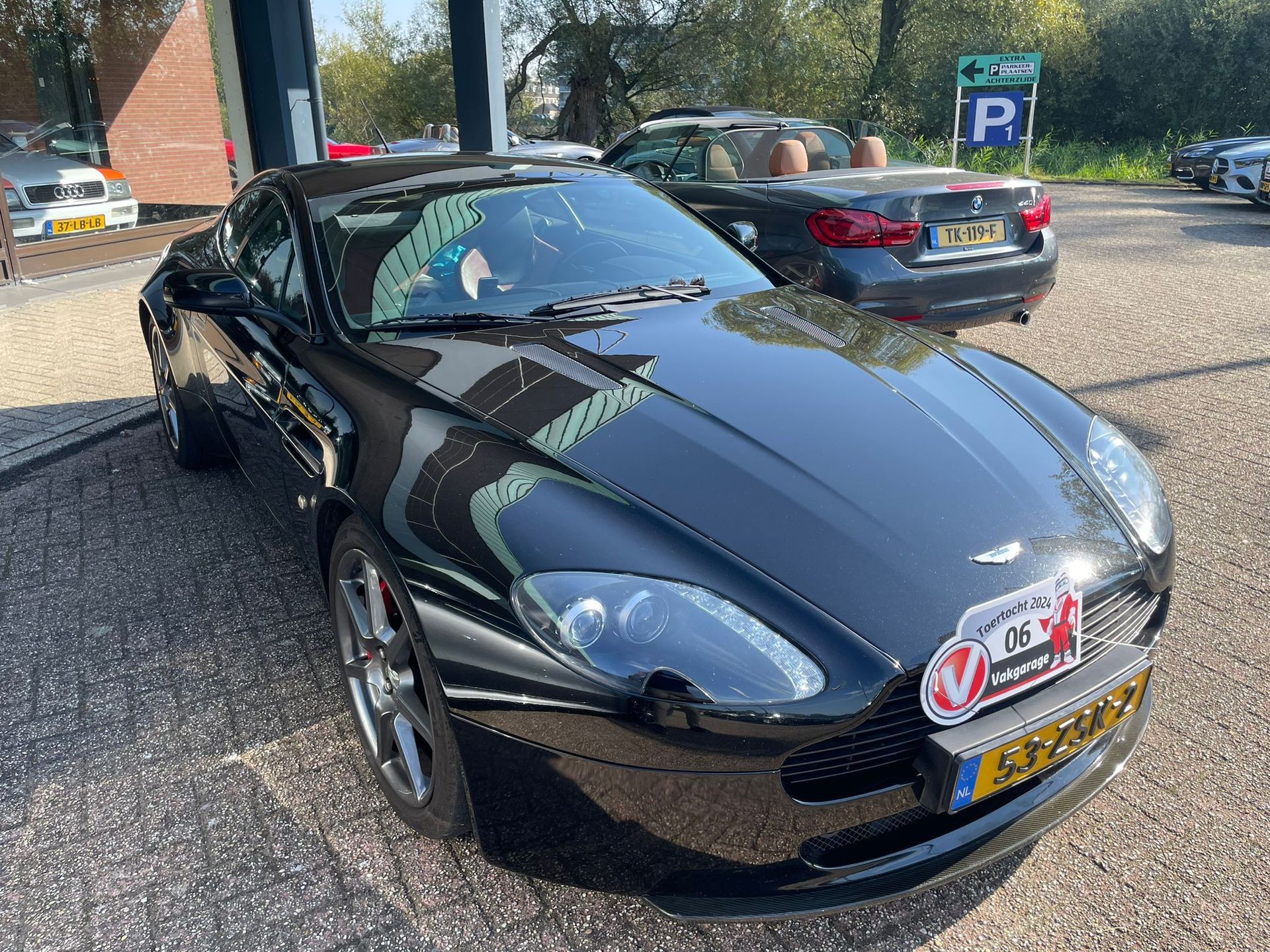 Een zwarte Aston Martin sportwagen staat geparkeerd voor een gebouw op een zonnige dag.