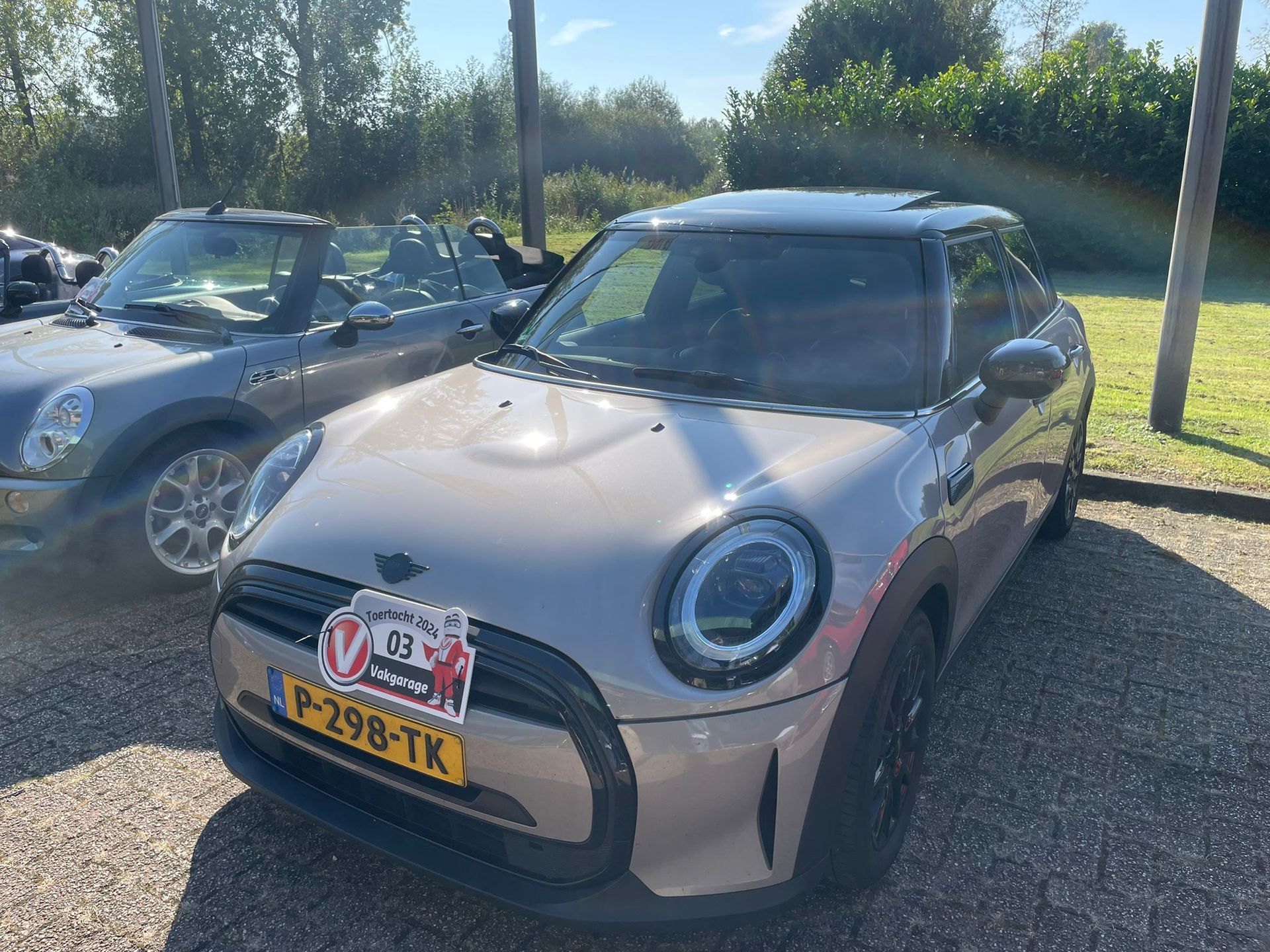 Een grijze Mini Cooper staat geparkeerd met op de achtergrond een andere zilveren Mini Cooper cabriolet.
