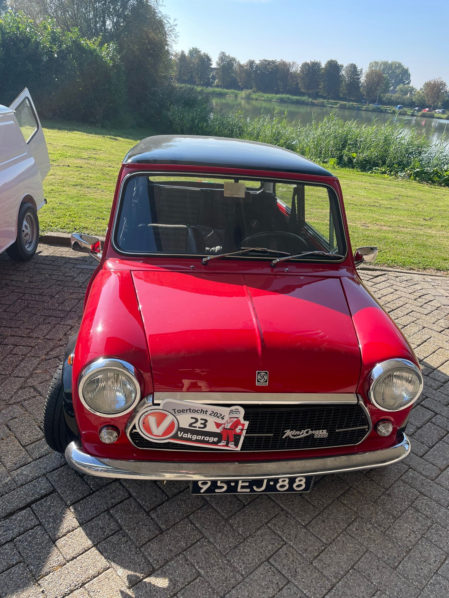Een rode klassieke Mini Cooper met zwart dak staat buiten geparkeerd, vlakbij het water.