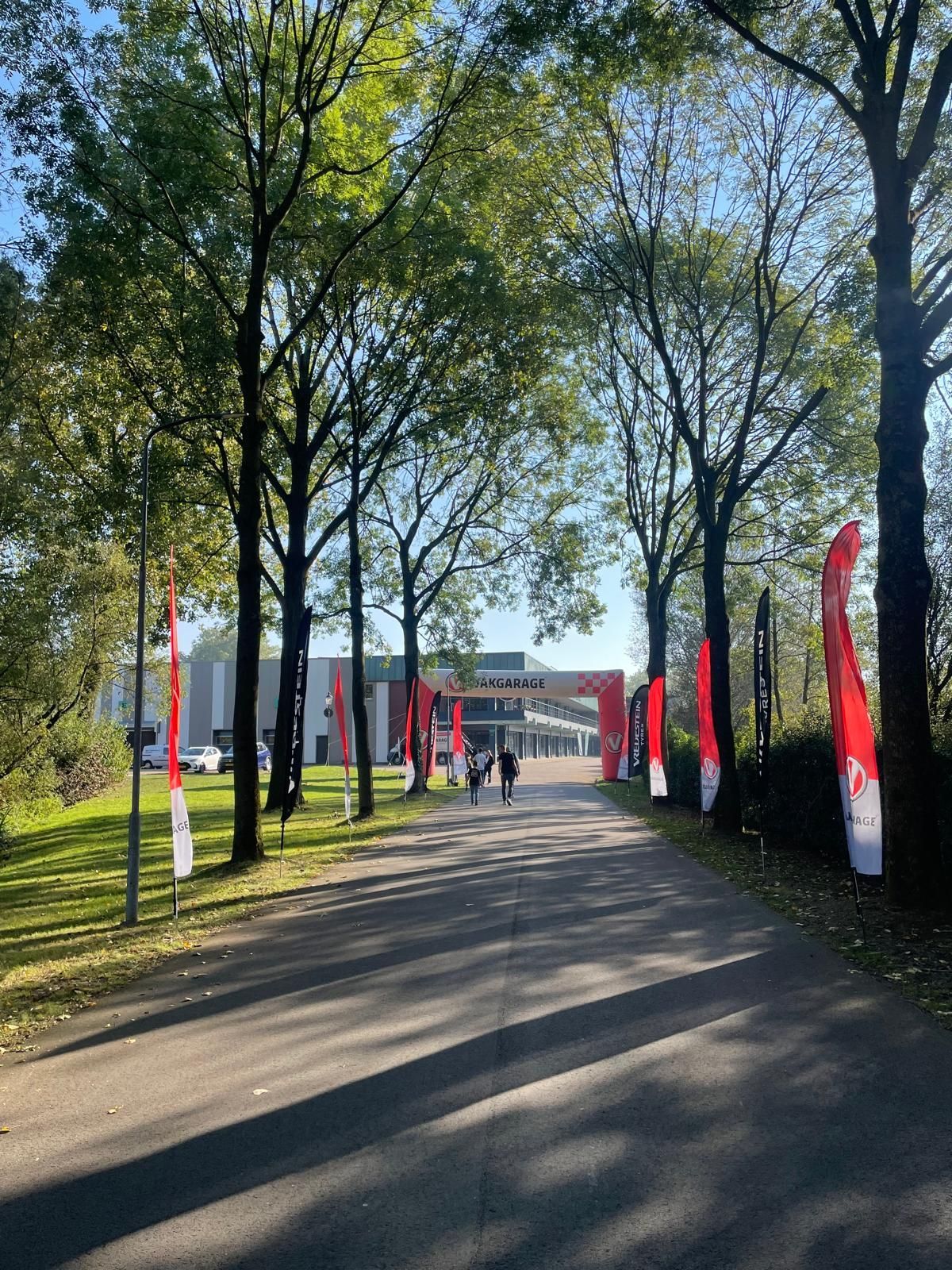 Een met bomen omzoomde weg die naar een gebouwingang leidt, met rode vlaggen en twee mensen die lopen.