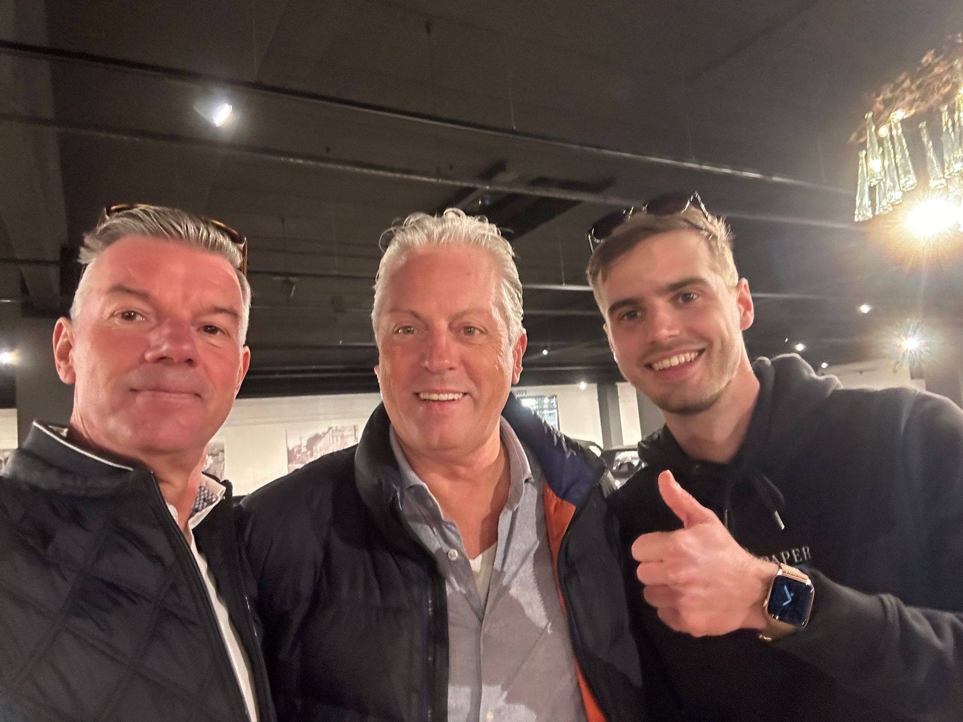 Drie mannen glimlachen, binnen. Een van hen steekt zijn duim omhoog.