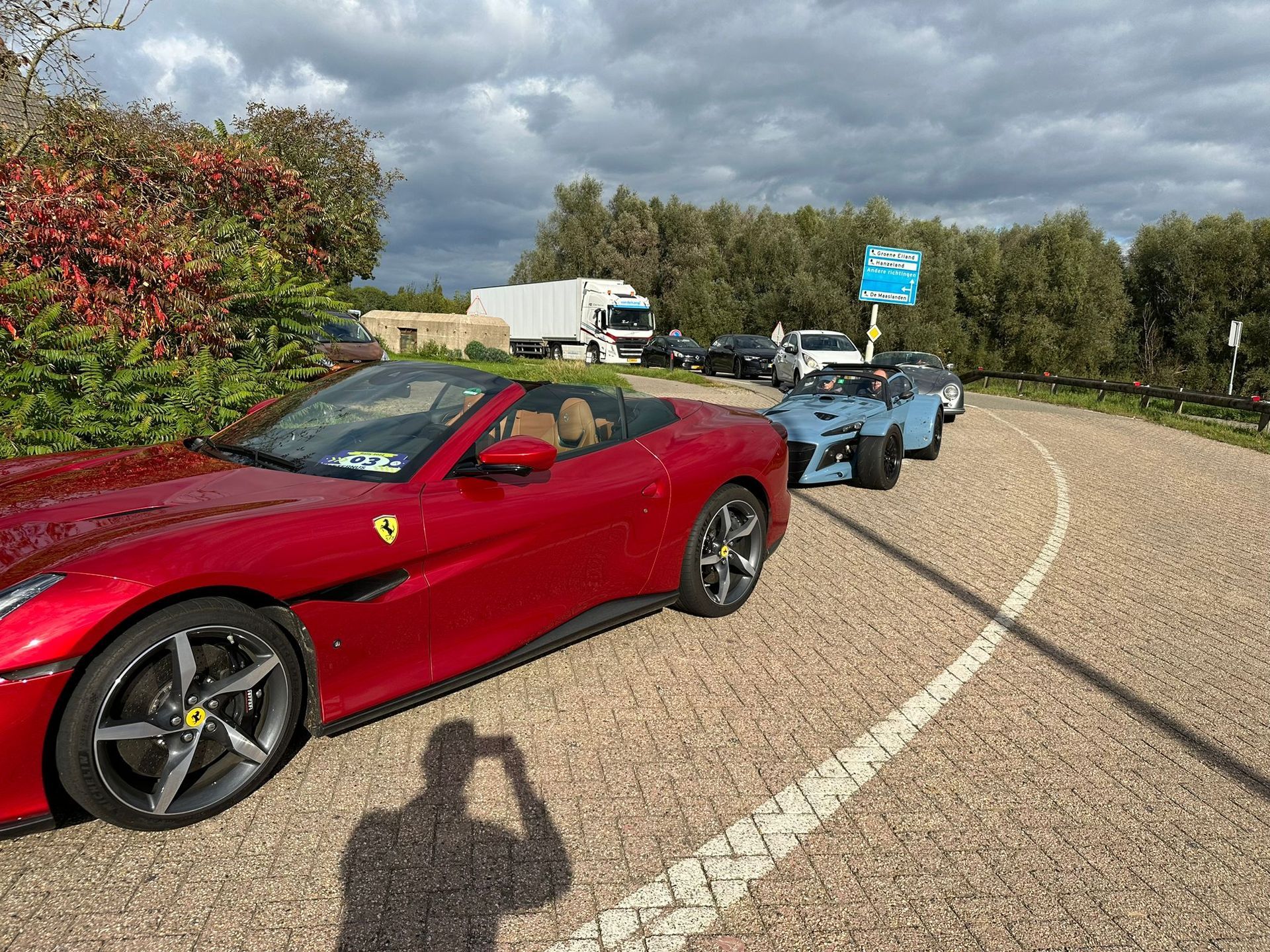 Een rode Ferrari cabriolet staat geparkeerd, gevolgd door andere auto's op een bochtige weg onder een bewolkte hemel.