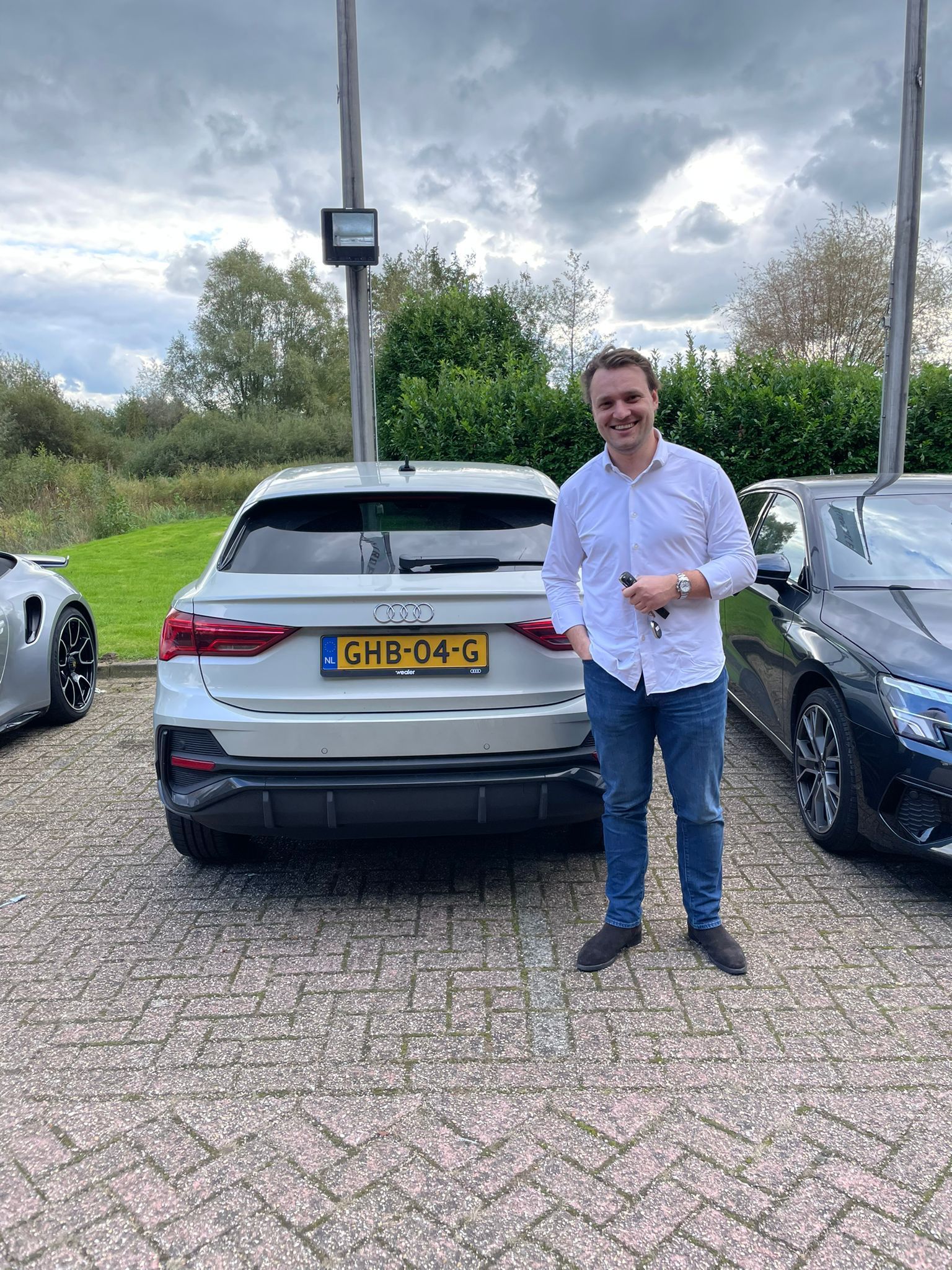 Een man staat naast een zilverkleurige auto met een Nederlands kenteken. Hij lacht in een buitenomgeving.