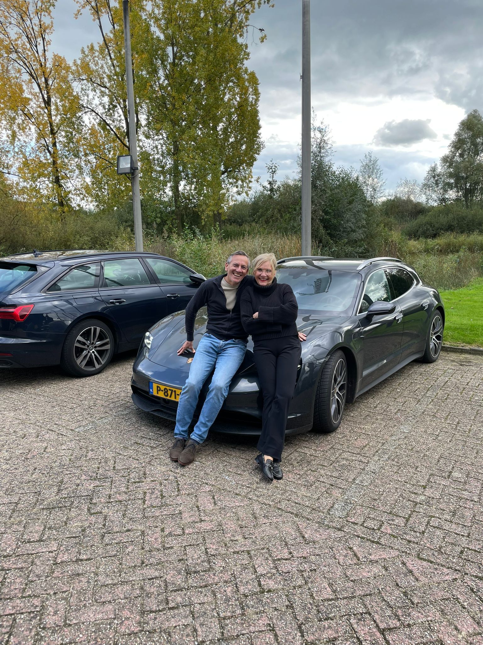 Twee mensen poseren op een donkere auto op een parkeerplaats op een bewolkte dag.