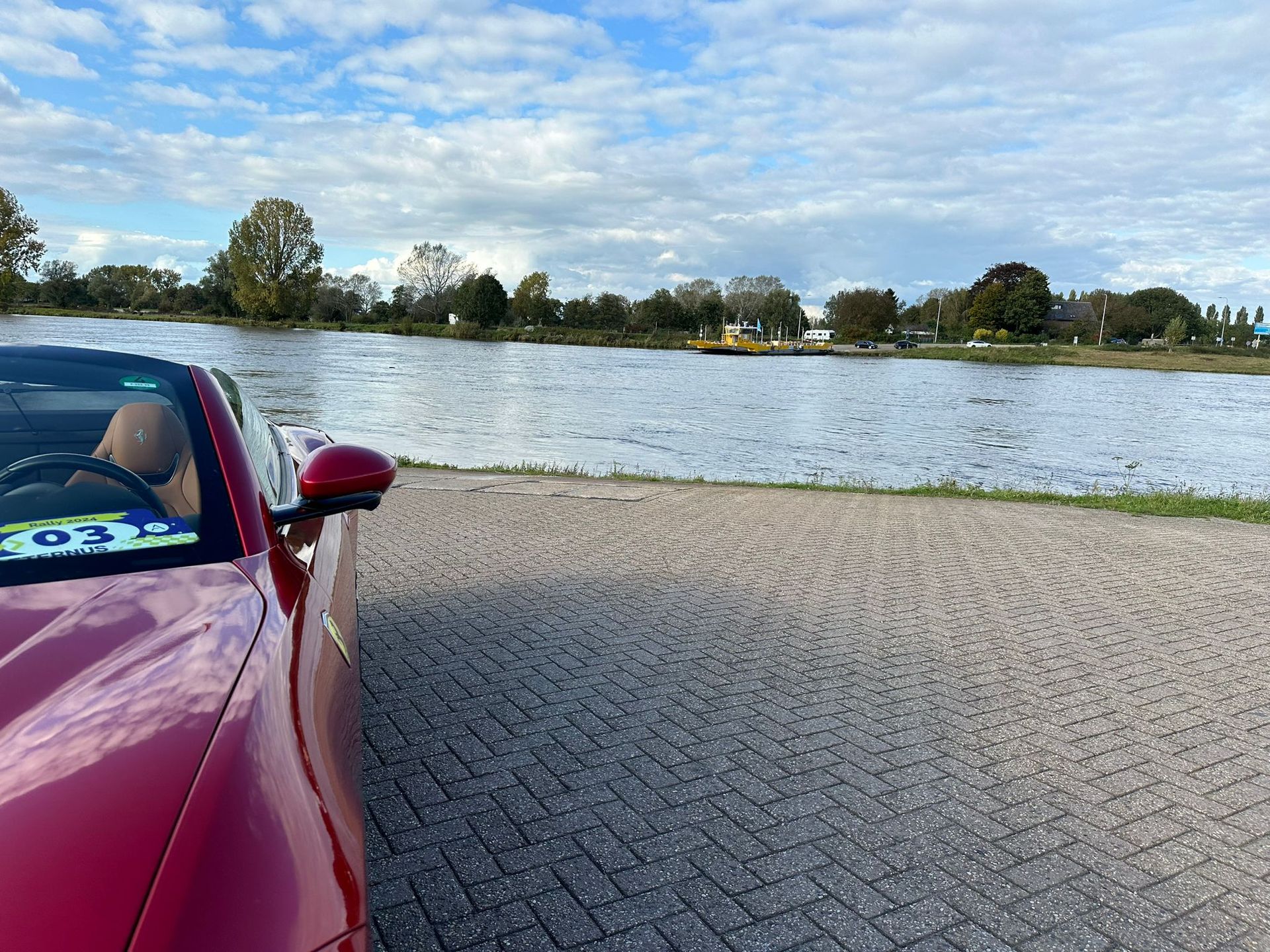 Een rode cabriolet staat geparkeerd naast een waterpartij onder een bewolkte hemel.