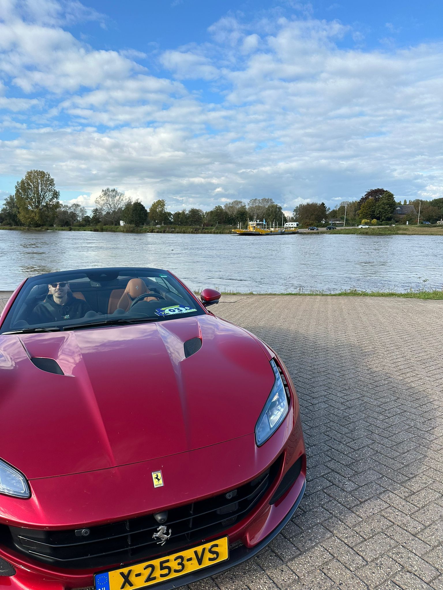 Een rode Ferrari cabriolet staat geparkeerd aan een meer onder een gedeeltelijk bewolkte hemel.
