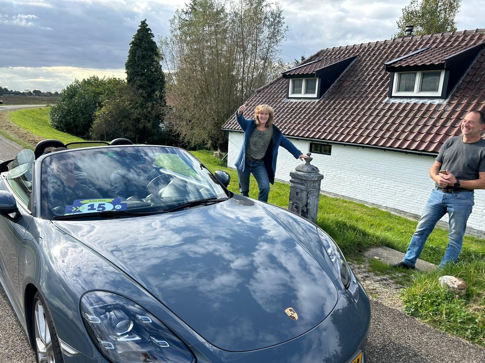 Grijze cabriolet met twee inzittenden langs een landweg. De ene persoon wijst, de andere gebaart. Op de achtergrond is een wit huis te zien.