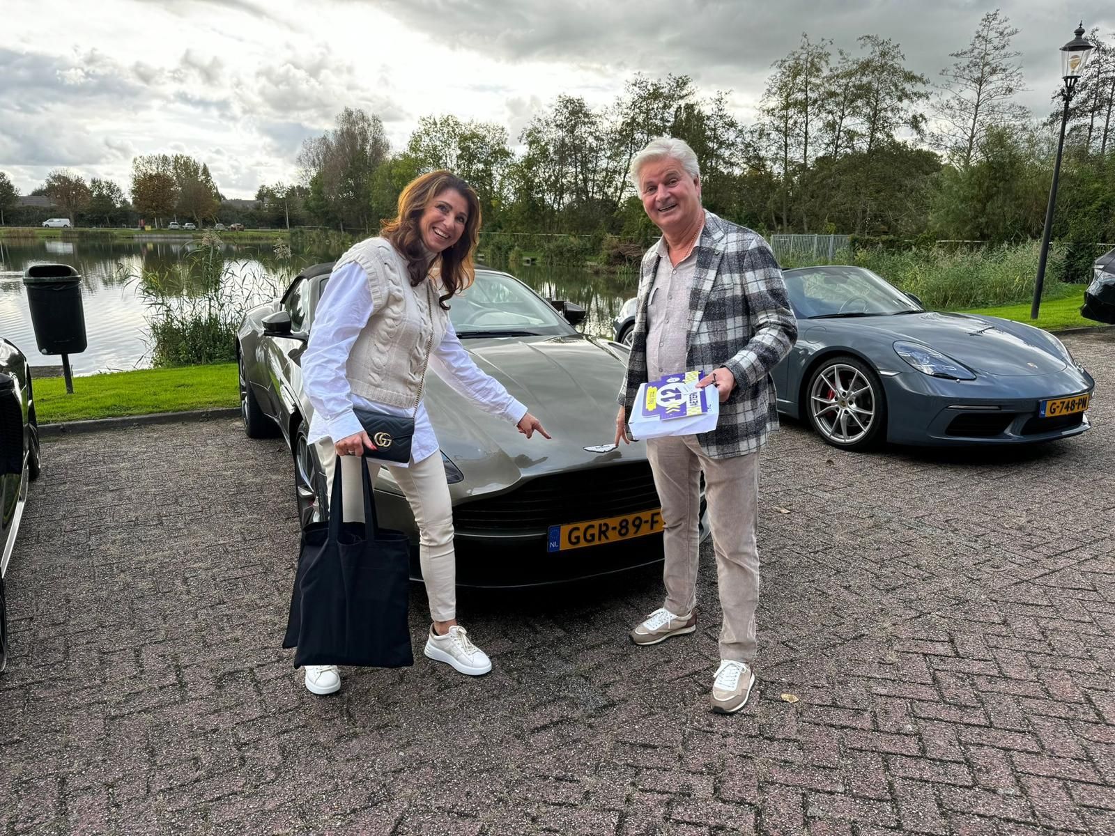 Een vrouw wijst naar een donkergrijze auto, een man houdt papieren vast. Ze staan ​​buiten bij het water, achter hen rijdt nog een auto.