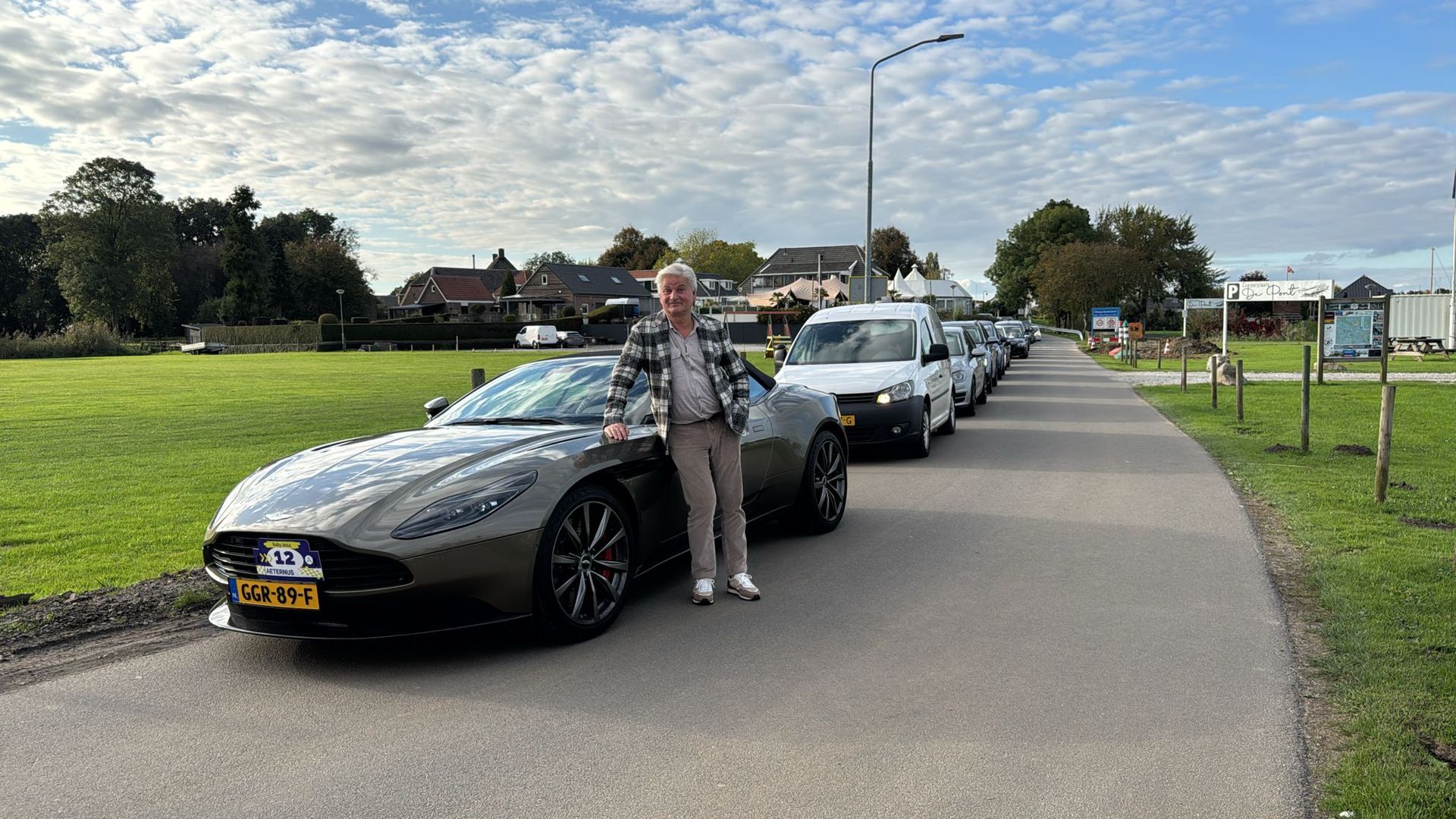 Een persoon leunt tegen een grijze sportwagen die geparkeerd staat op een weg met andere voertuigen aan weerszijden en een grasveld op de achtergrond.